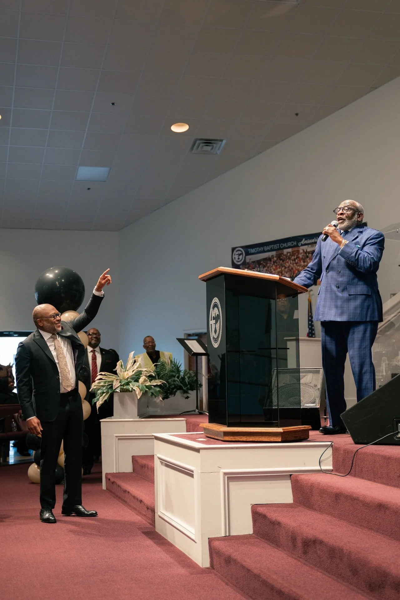 A man in a blue plaid suit speaking at a church podium with a microphone, while another man in a dark suit points towards him. There are people in the background and decorations on the stage, including balloons and plants.
