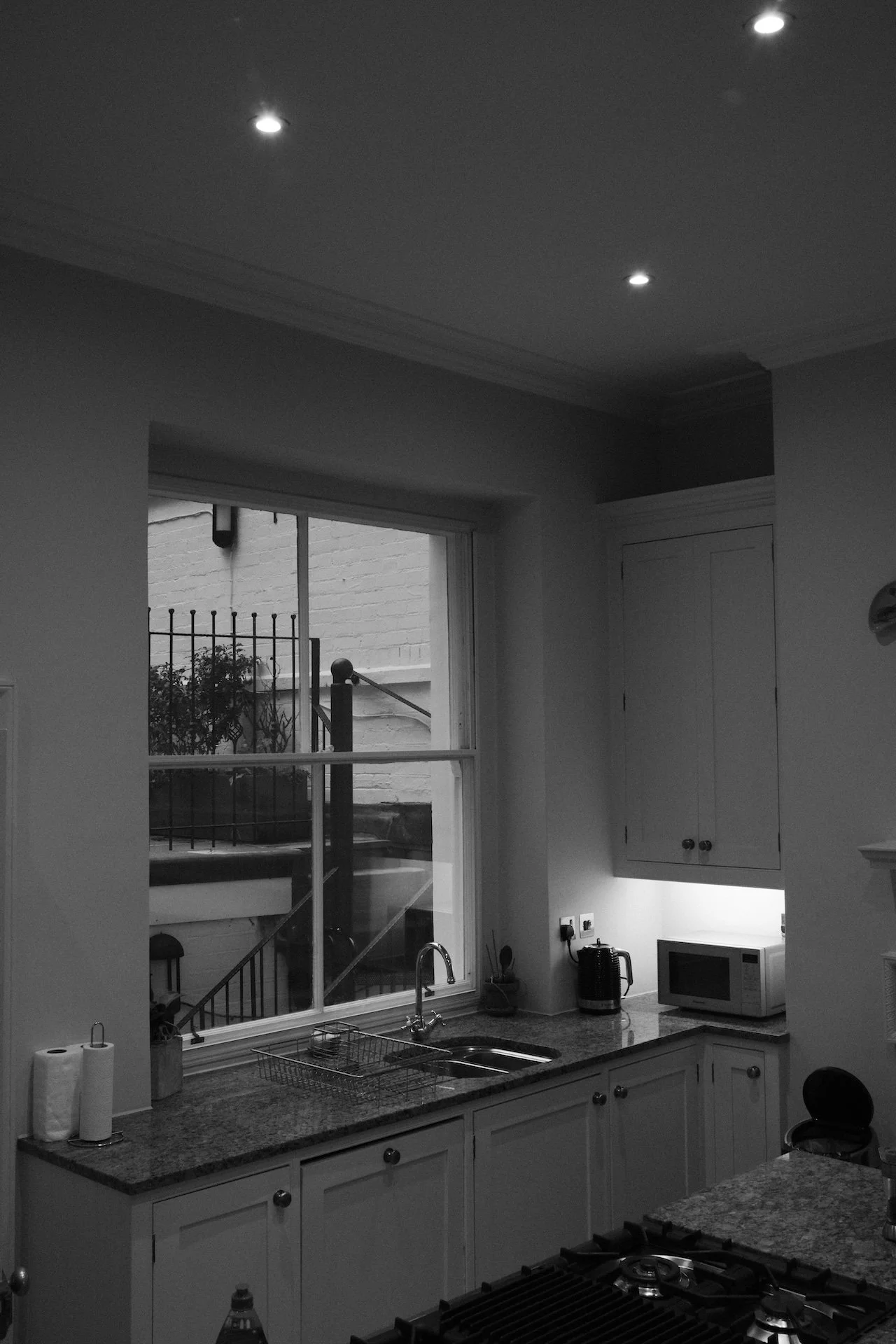 A black and white photo of a kitchen with a window showing an outside staircase, a countertop with a sink, kettle, microwave, and paper towel roll, and ceiling lights.