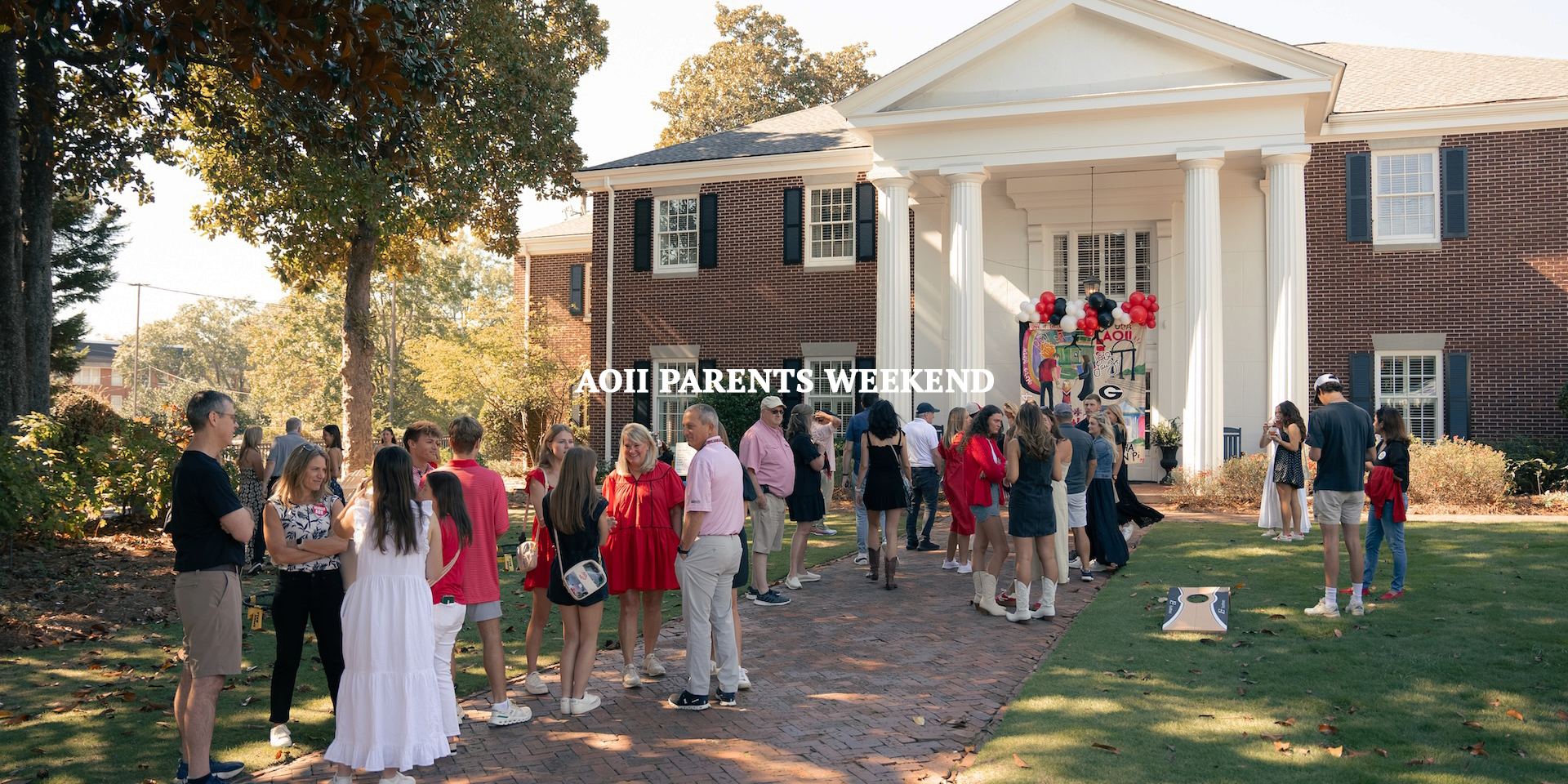 A group of people gathered outside a large brick and white house for an event called AOII Parents Weekend, with balloons and decorations on the house's front porch.