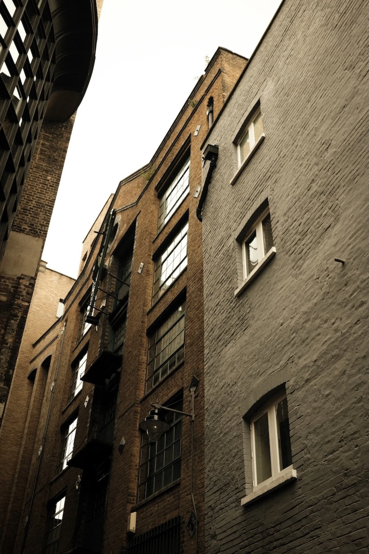 Low-angle view of two adjacent brick buildings with large windows, fire escapes, and street lamps, under a cloudy sky.