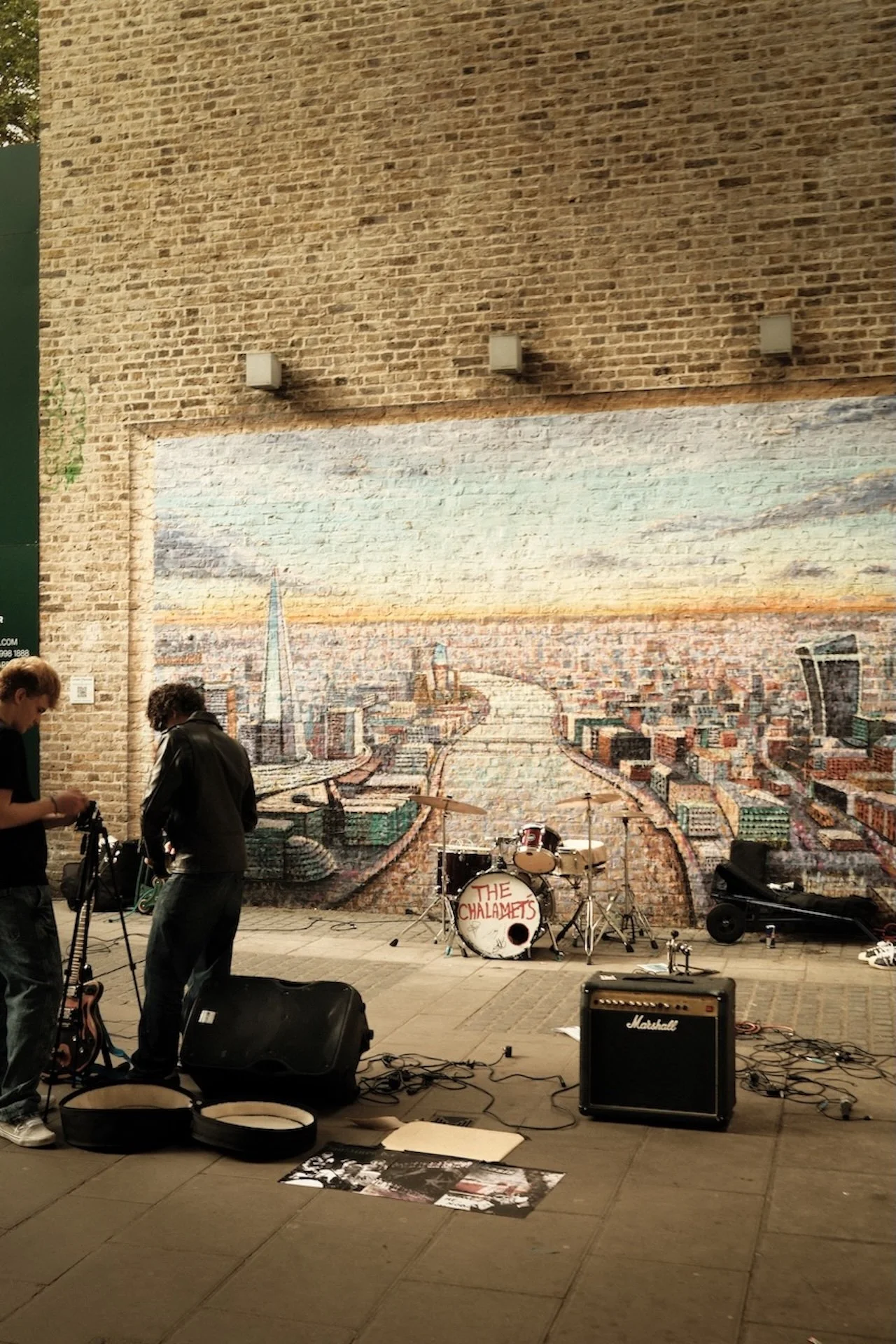 Street scene with a large brick wall featuring a mural of a cityscape, a drum set labeled 'The Chalamets,' and two men setting up musical equipment.
