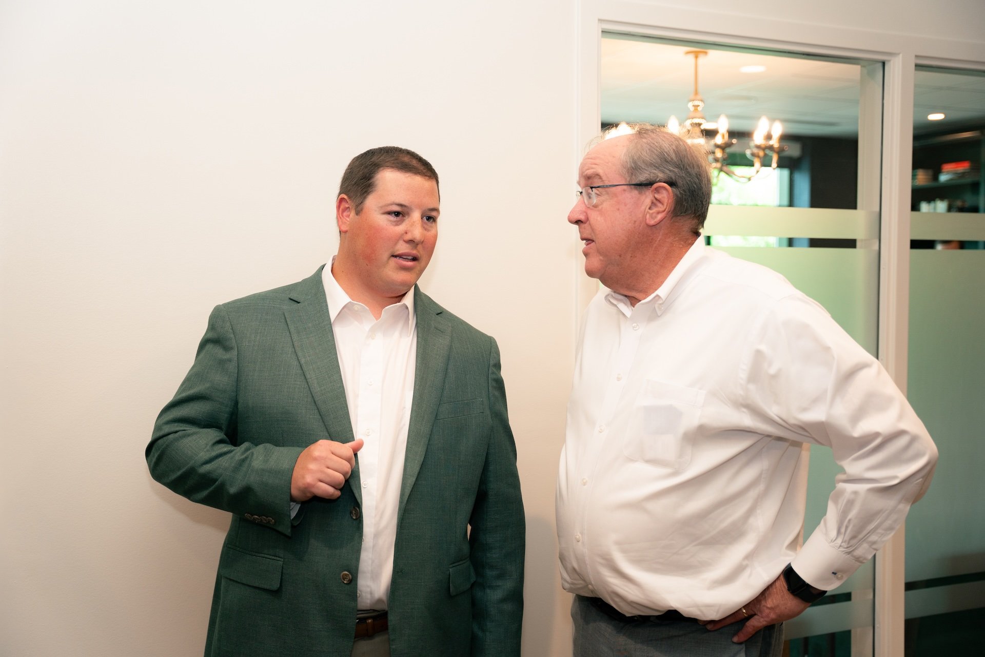 Two men are having a conversation indoors, one younger in a green suit and white shirt, and one older in a white shirt. There is a glass window behind them and a chandelier is visible in the background.