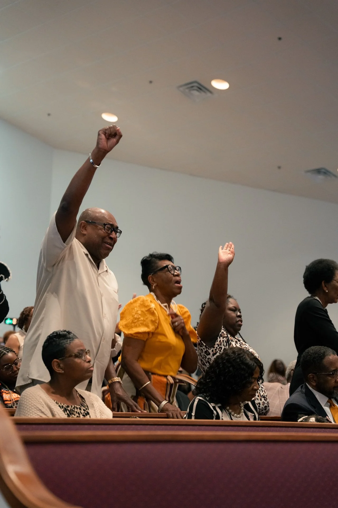 People standing and raising their hands in a congregation or service, with some seated in the foreground.