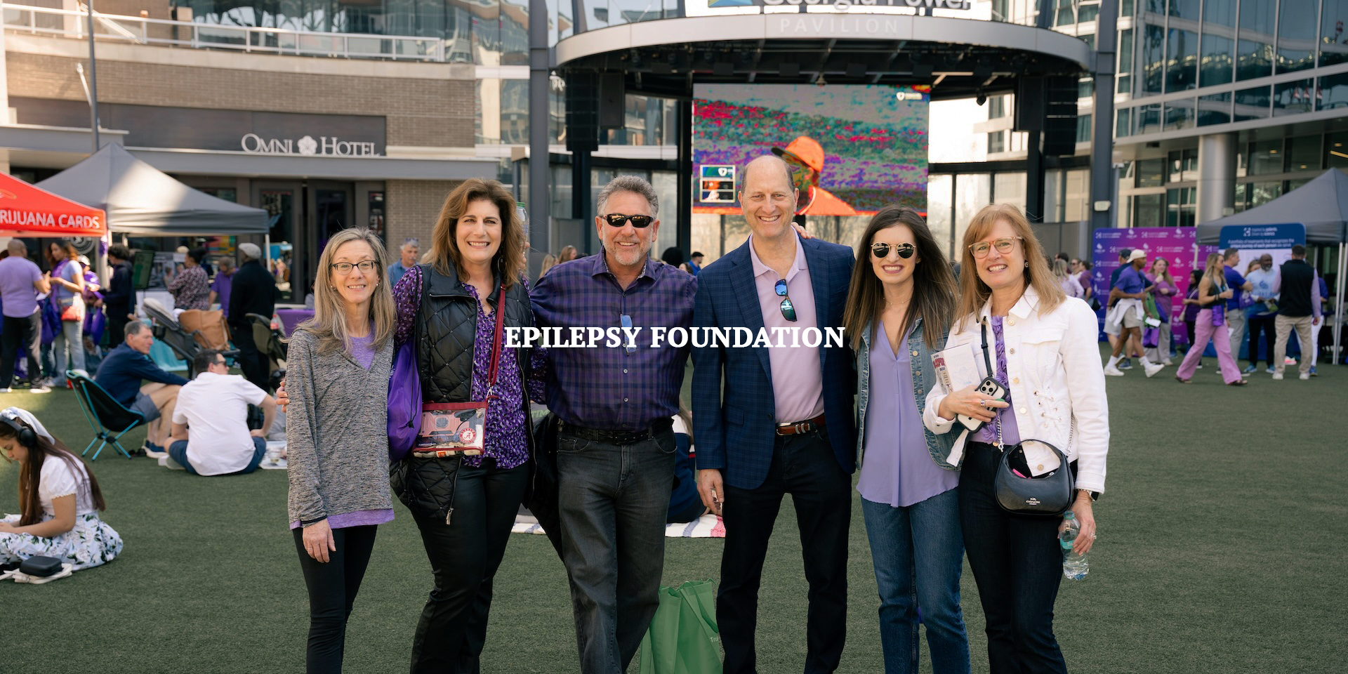 Group of six people posing and smiling at an outdoor event during daytime, with tents, booths, and people in the background. The event is associated with the Epilepsy Foundation.