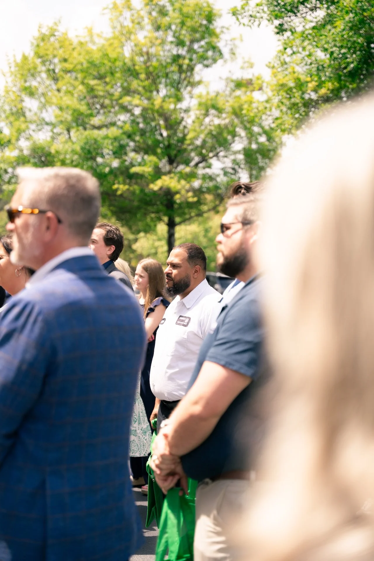 People standing outdoors, attending a formal event or ceremony, with some holding green tote bags, and trees in the background.