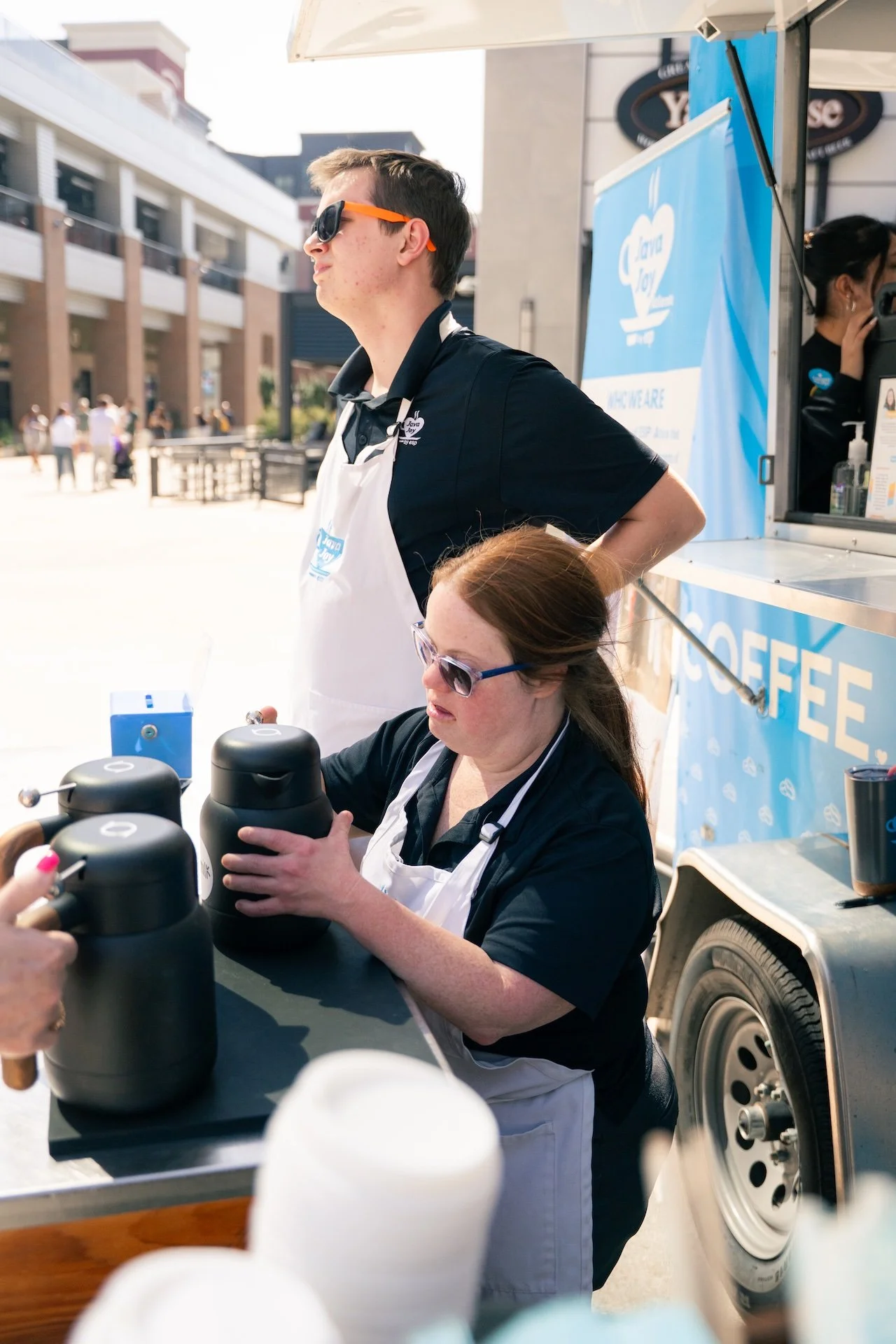 Two women working at a coffee stand outdoors, one is standing with sunglasses, the other is pouring coffee from a pot, with a blue coffee-themed banner in the background.
