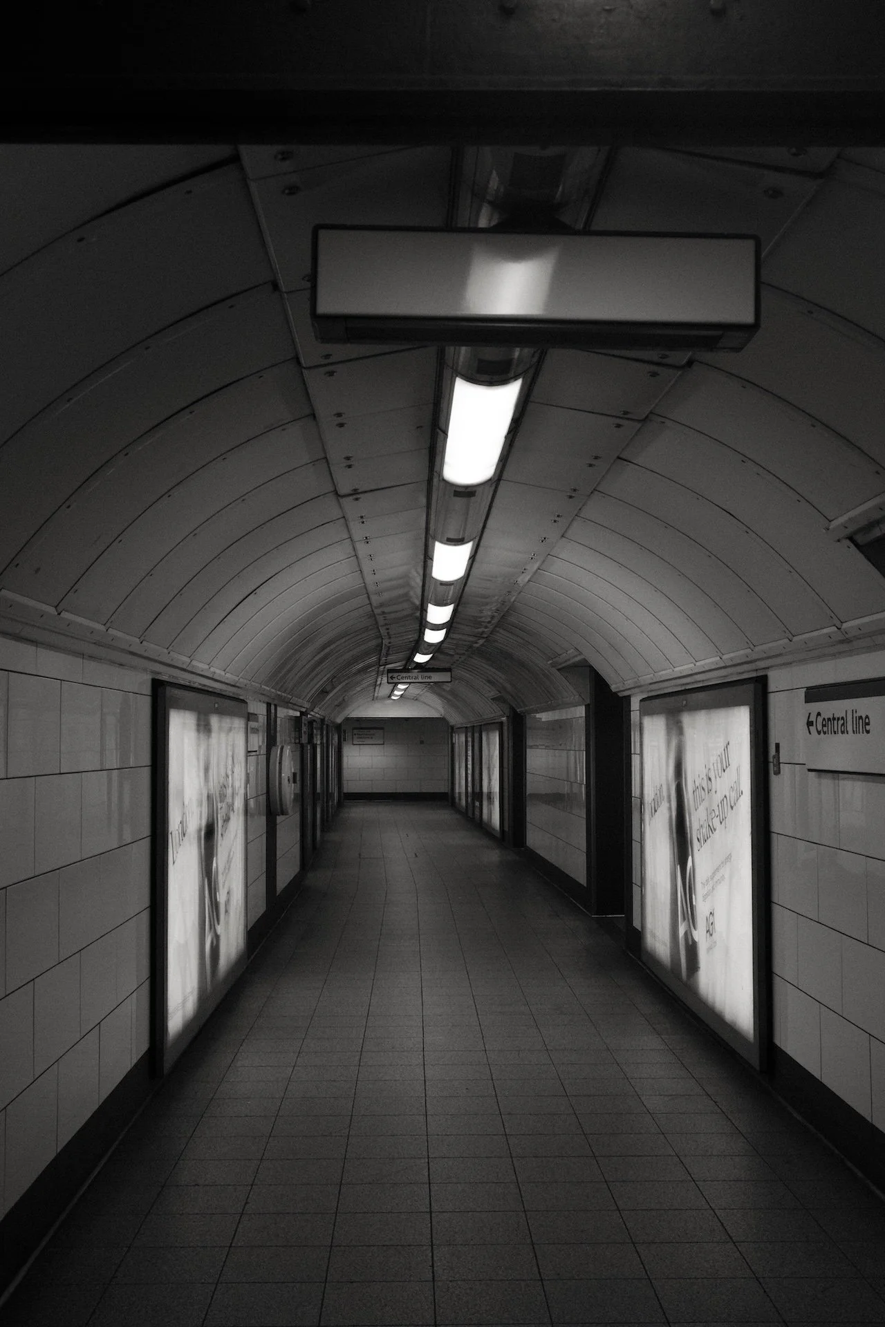 Underground subway tunnel with a curved ceiling, fluorescent lights, and advertising panels on the walls.