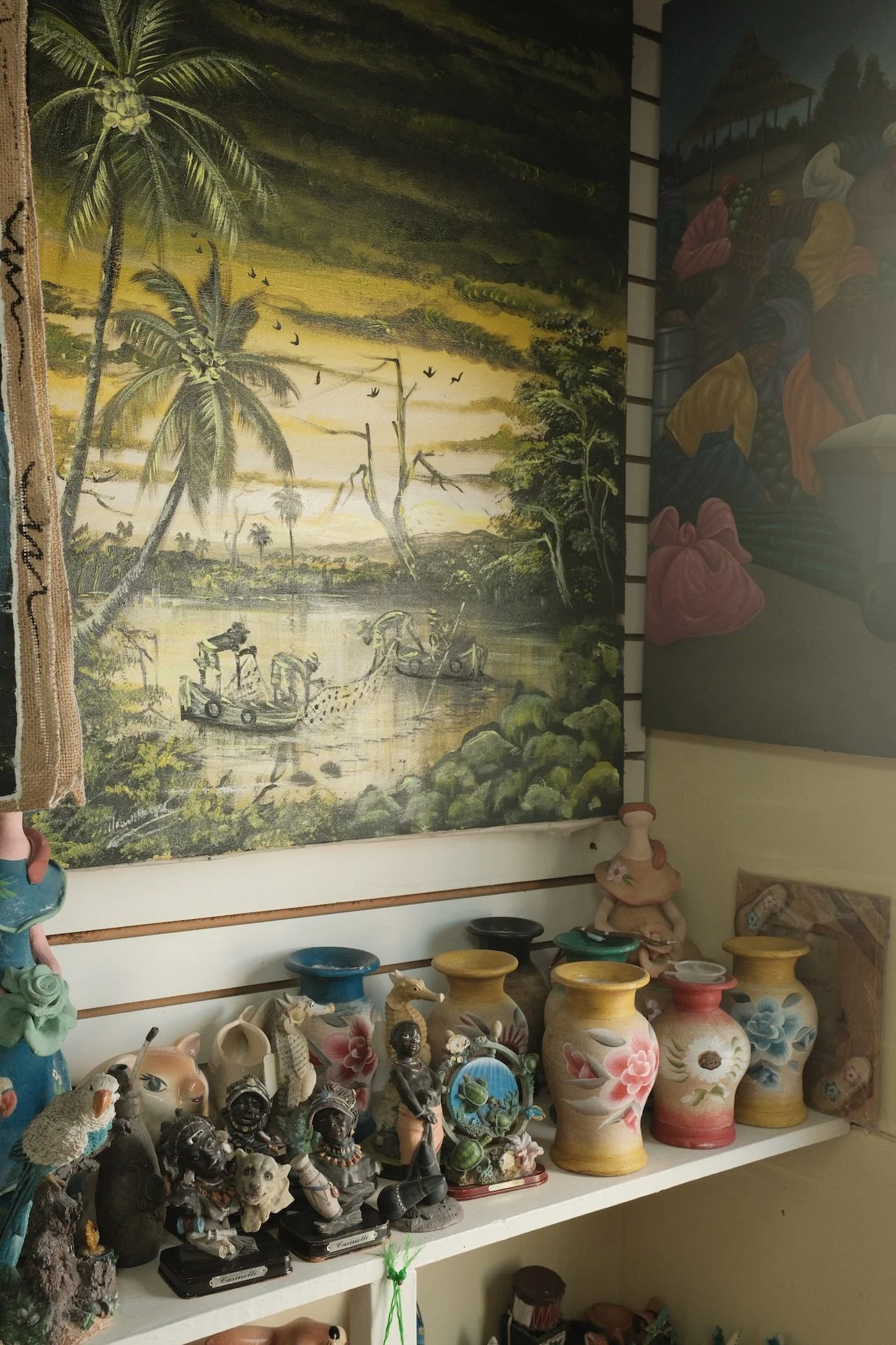 A store display with a large painting of a tropical landscape featuring palm trees, birds, and water, and a shelf below holding various decorative ceramics and figurines.