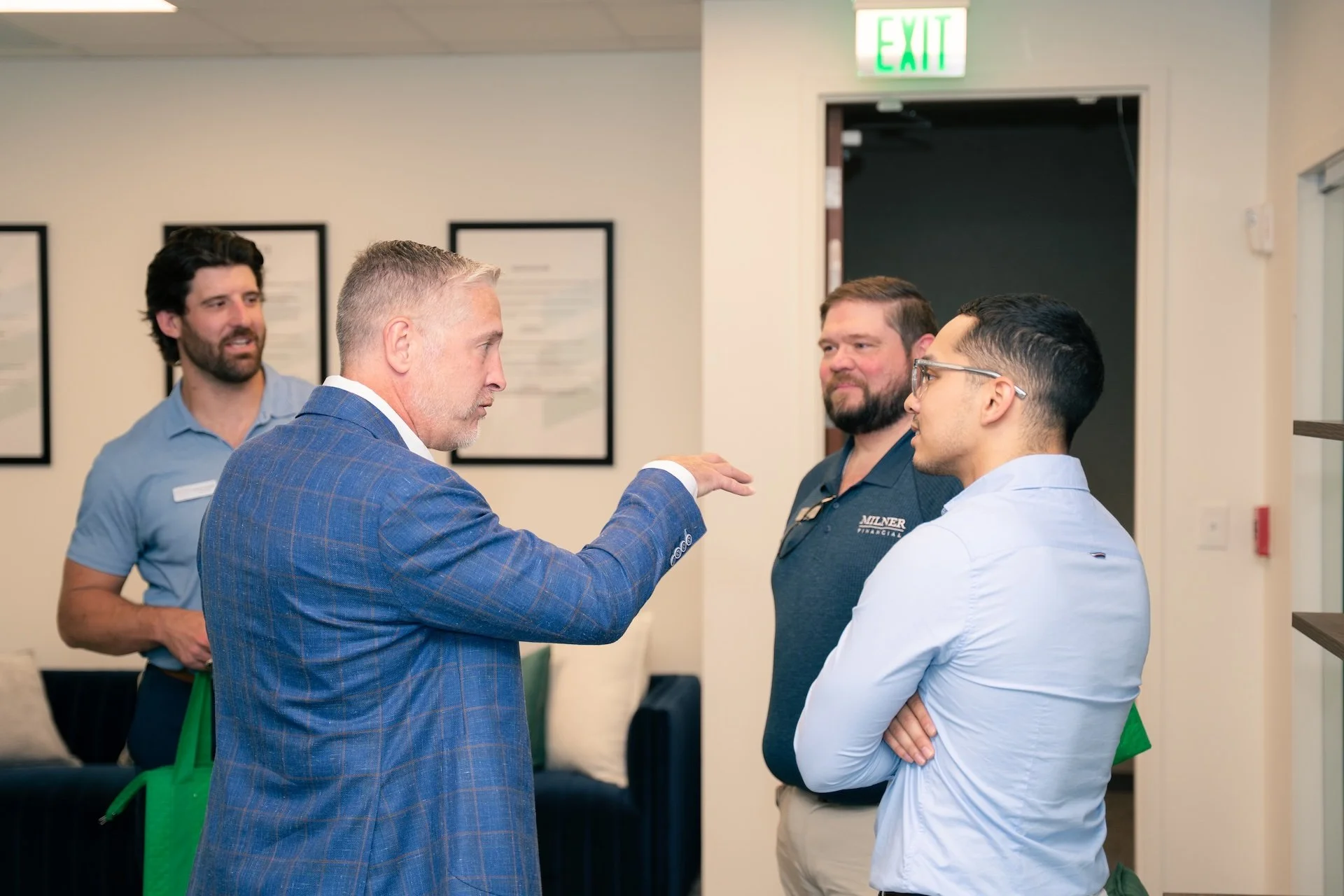Four men engaged in conversation in an indoor setting, with artwork on the wall and an illuminated "EXIT" sign above a doorway in the background.