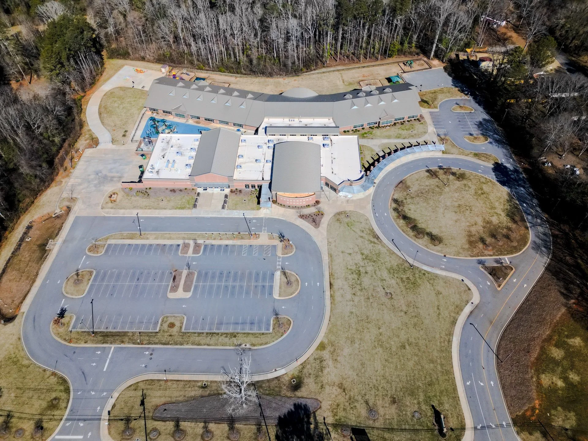 An aerial view of a school campus with a parking lot, playground, sports fields, and a surrounding wooded area.