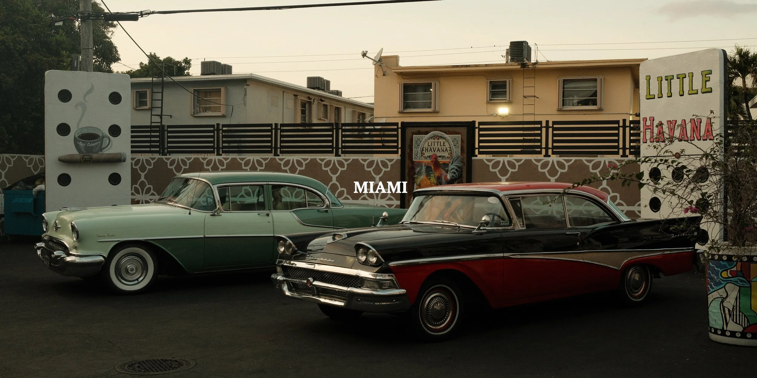 Two vintage cars, one black and red and the other green and cream, parked in front of a Miami restaurant named Little Havana with colorful signs and murals.