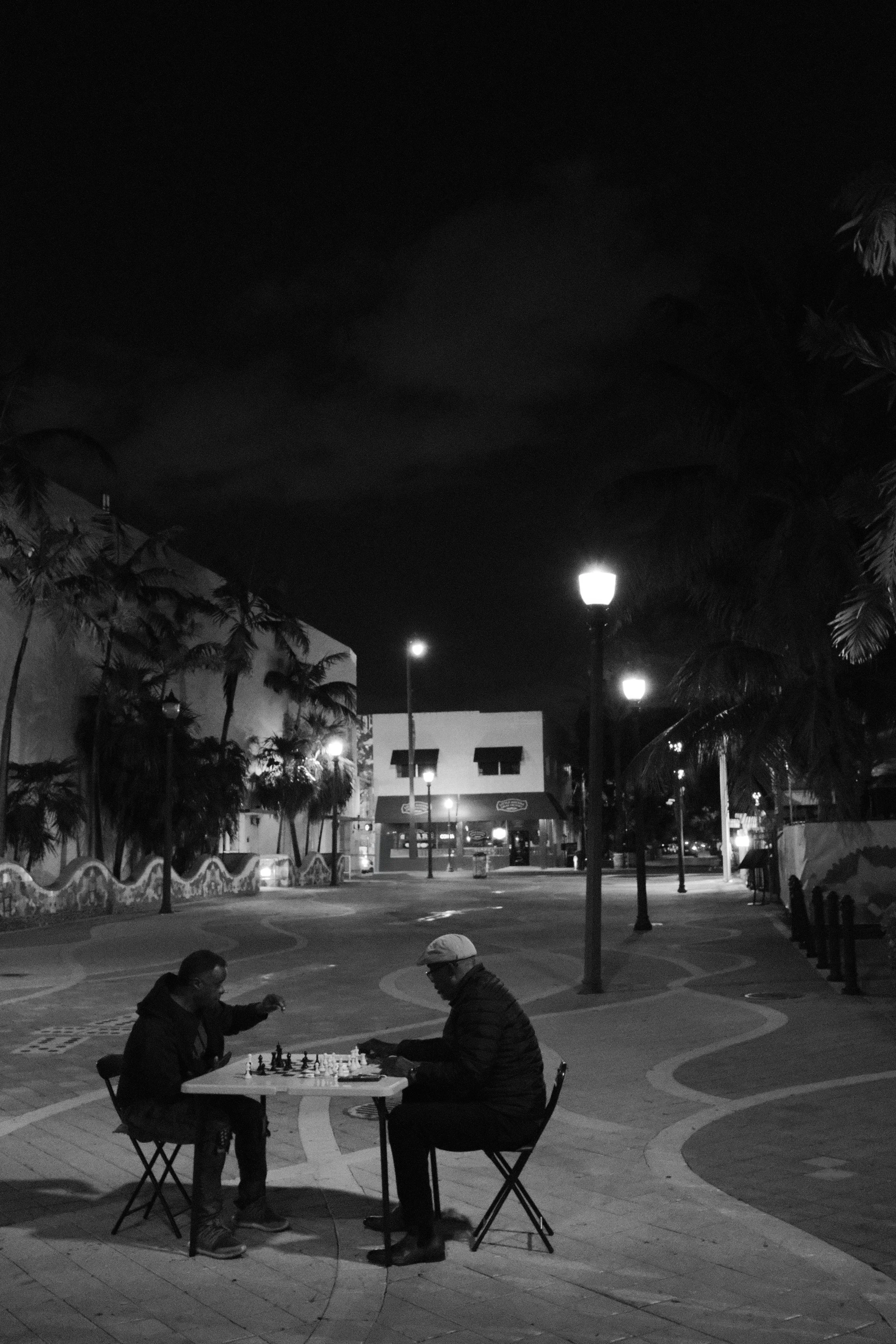 Two people playing chess at a table outdoors at night, illuminated by street lamps with a dark sky and buildings in the background.