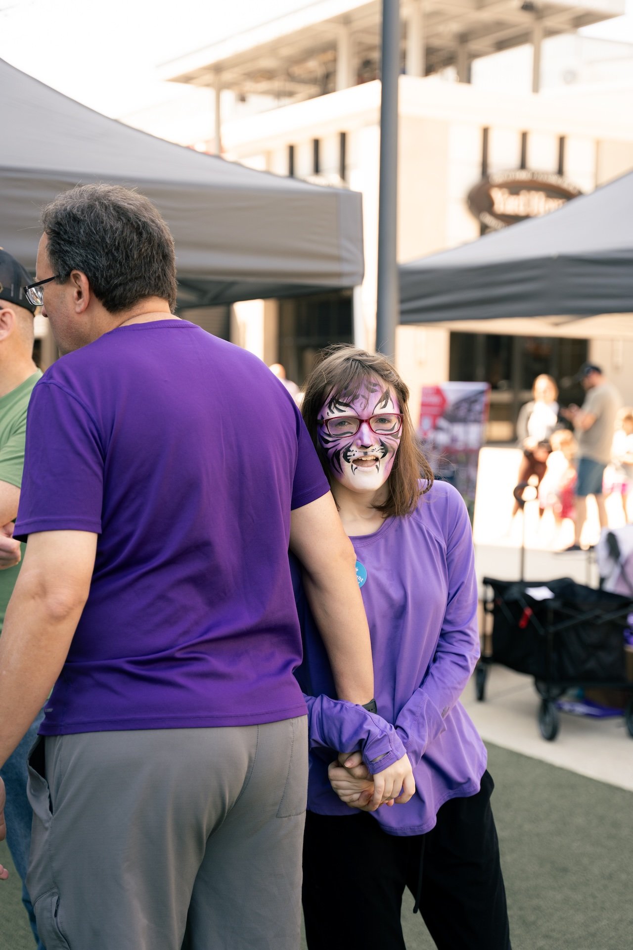 A woman with face paint resembling a tiger and wearing glasses, smiling and standing behind a man in a purple shirt at an outdoor event.