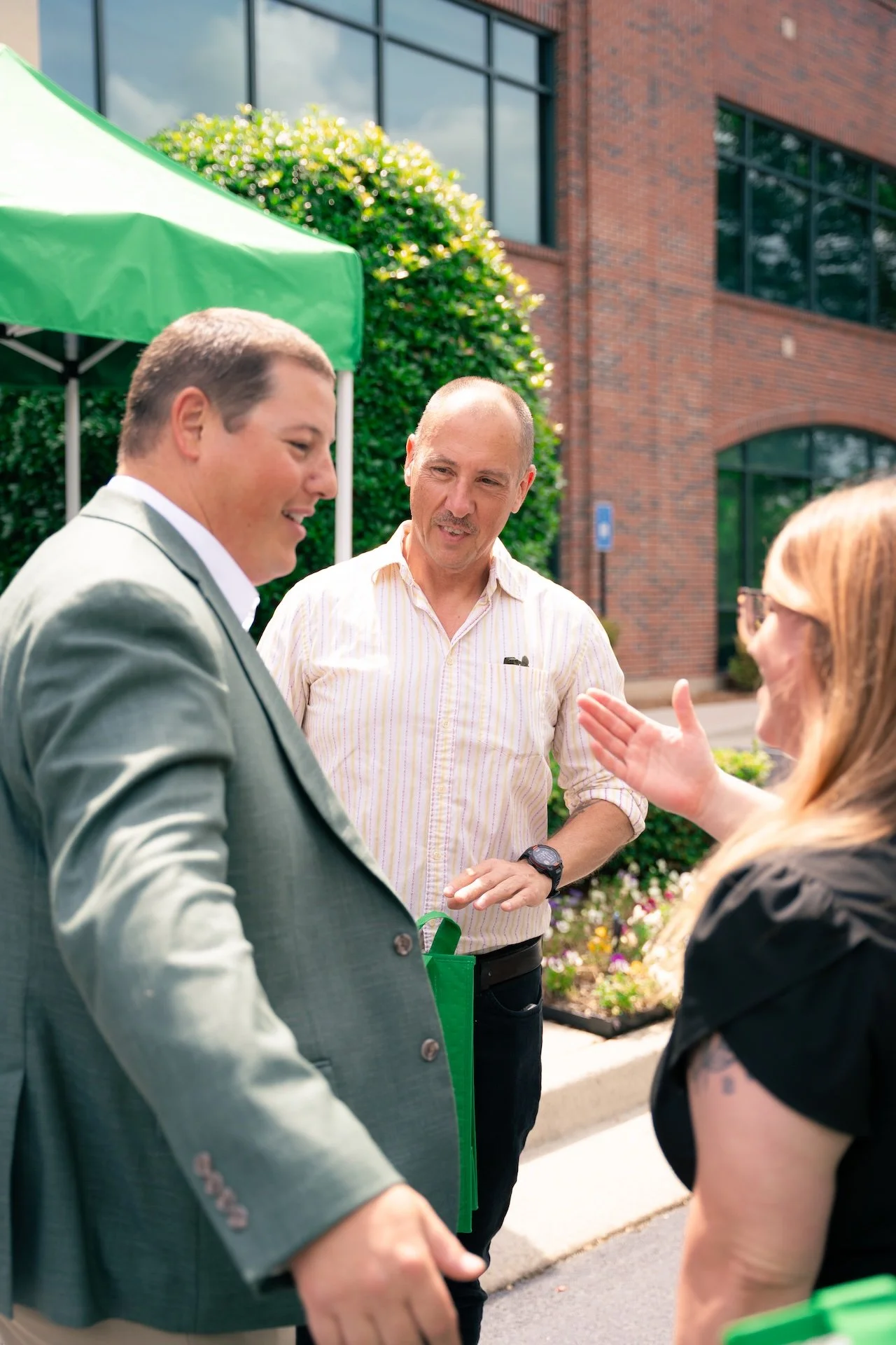 Three people engaged in conversation outdoors in front of a brick building and greenery, with a green tent canopy visible in the background.