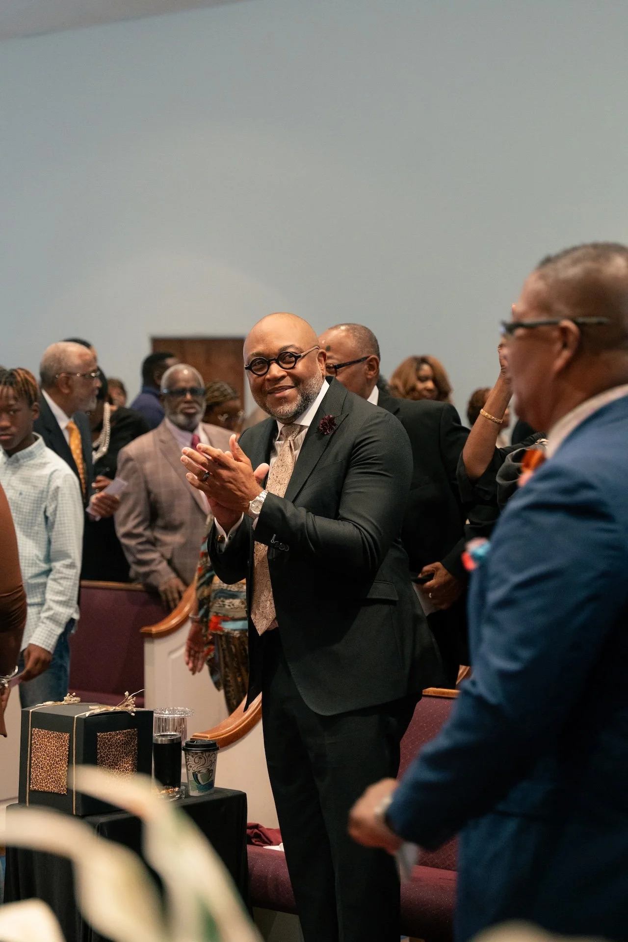 A man in a dark suit with glasses is clapping and smiling at a gathering of formally dressed people in a church or auditorium setting.