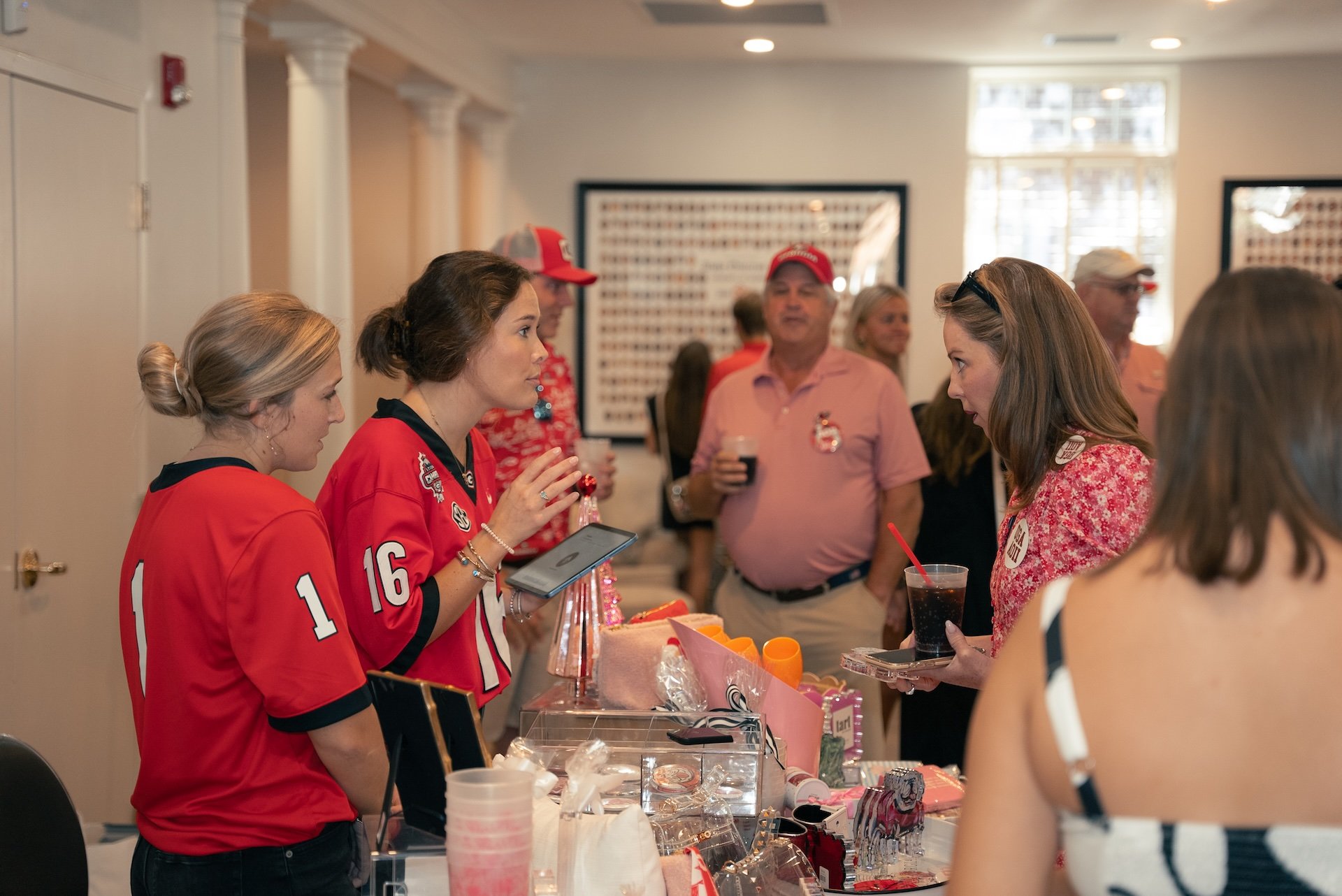 People at a social event, with women in sports jerseys and women holding drinks, standing around a table with various items.