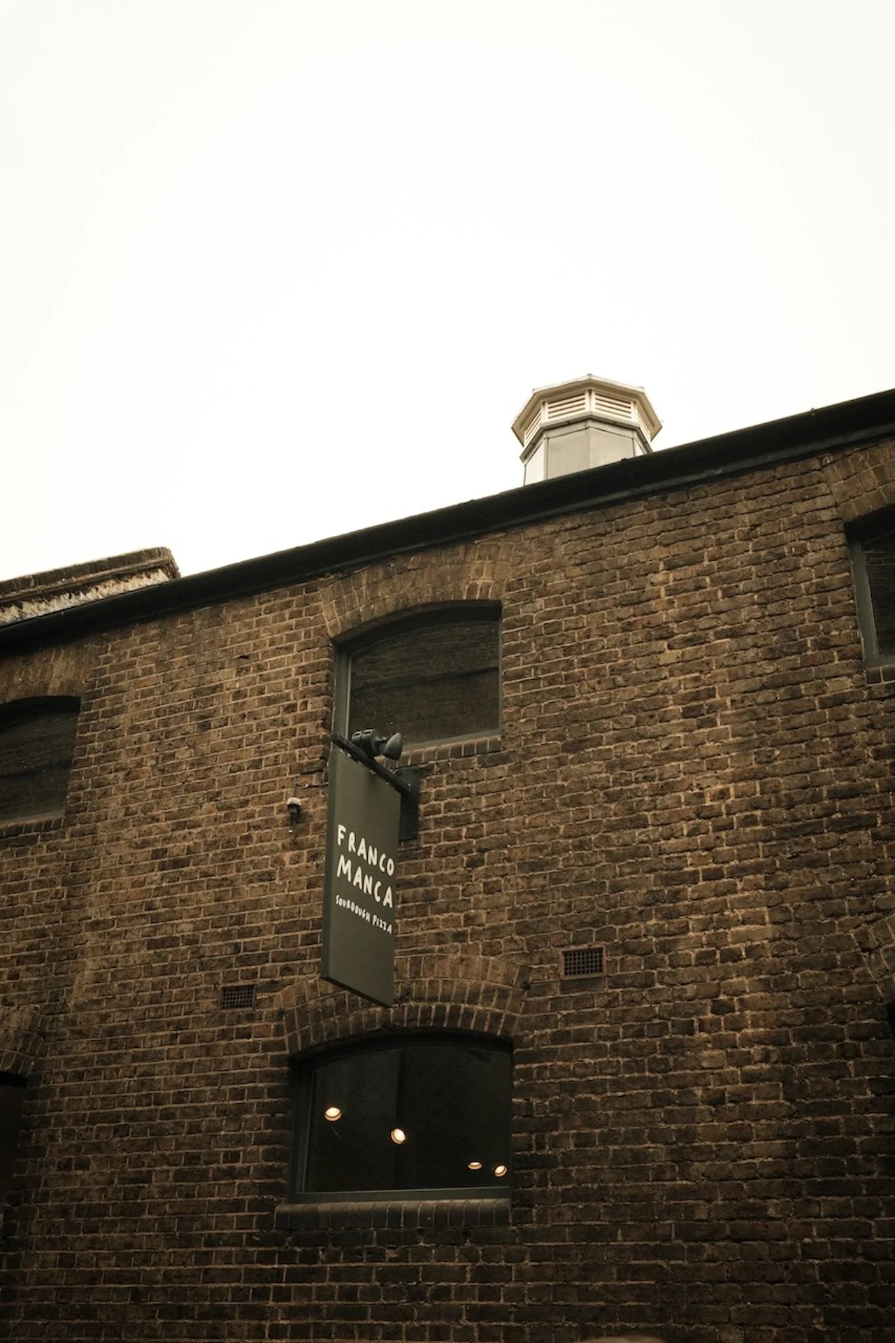 Exterior view of a brick building with a sign that reads 'Franco Manca Soufood Pizza,' and a vent on the roof against a cloudy sky.