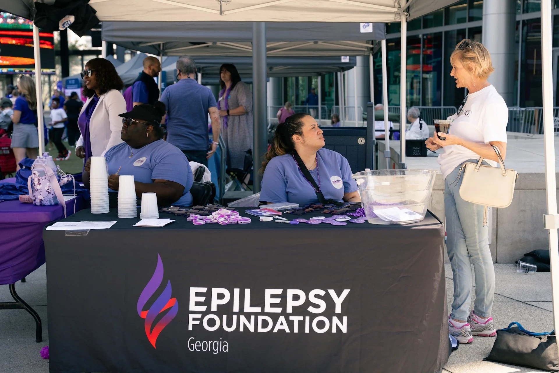 A woman in a white t-shirt and light blue pants standing at a booth with a black tablecloth that reads 'Epilepsy Foundation Georgia.' The booth has purple and pink items on display, and two women in purple shirts sit behind the table. There are sever