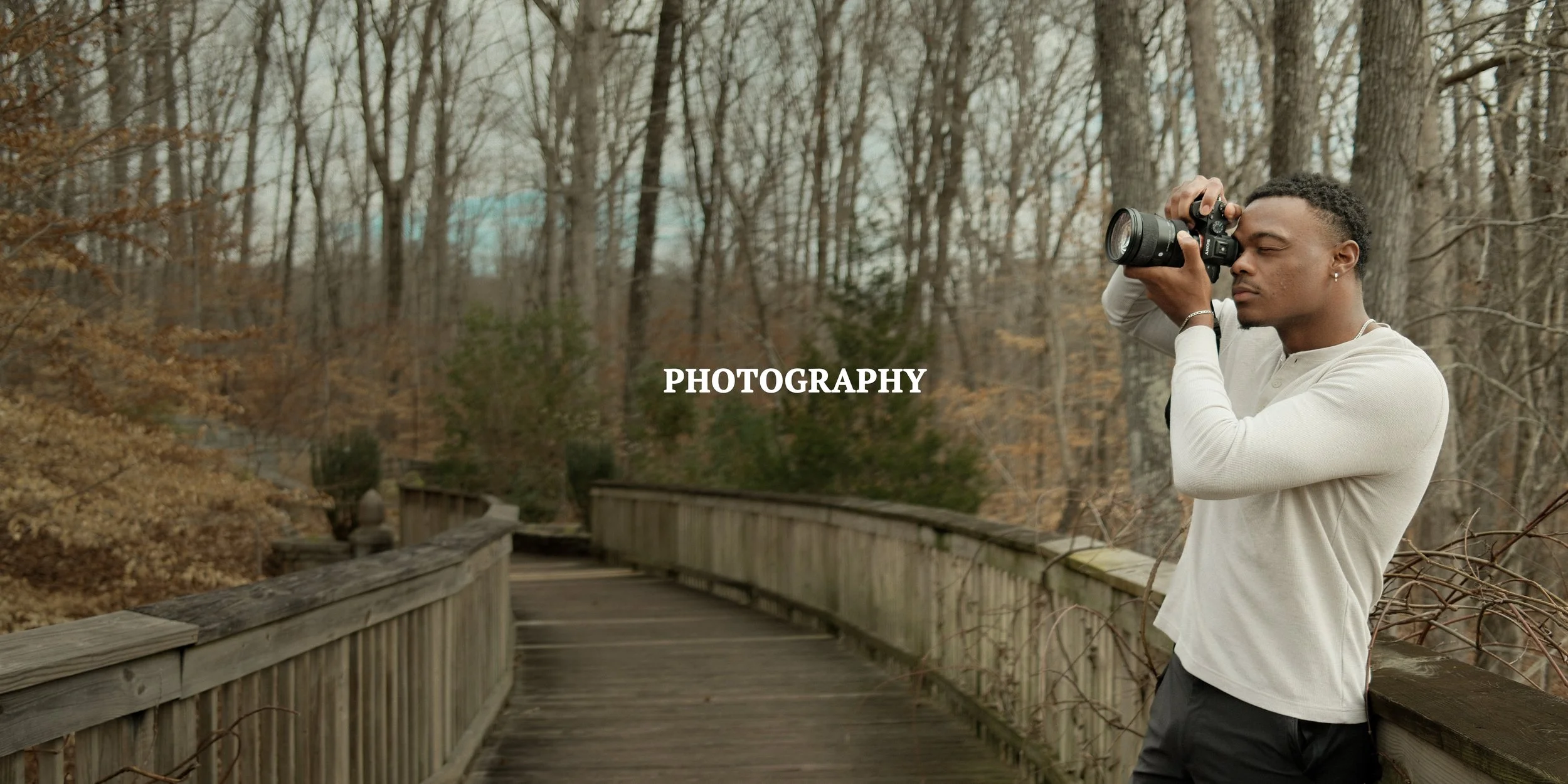 A man is standing on a wooden bridge in a forest, holding a camera up to his face and taking a photo. The trees in the background have no leaves, suggesting it's late fall or winter. The scene appears to be overcast.