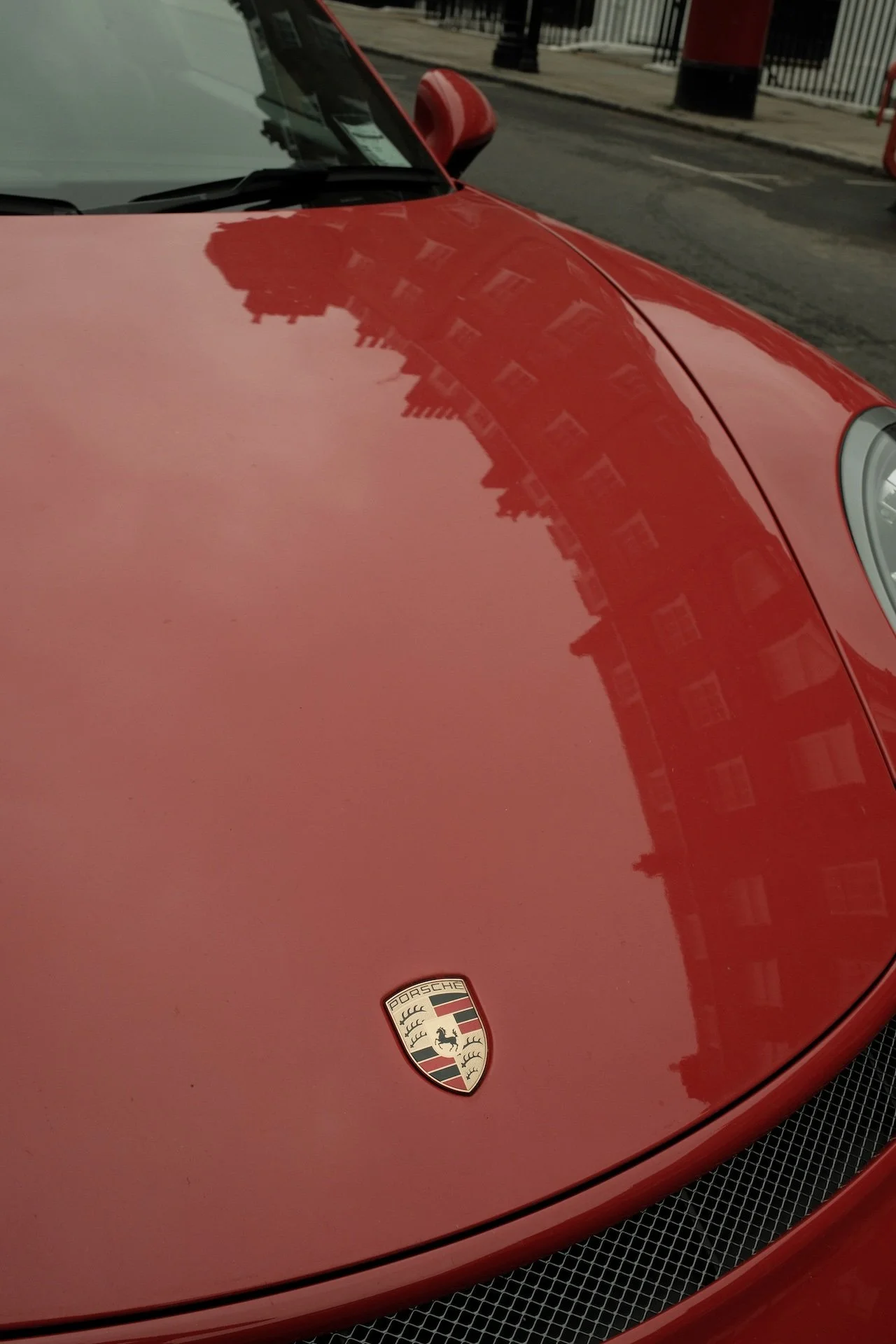 Close-up of a red Porsche car hood with a Porsche emblem, reflecting nearby buildings and street surroundings.