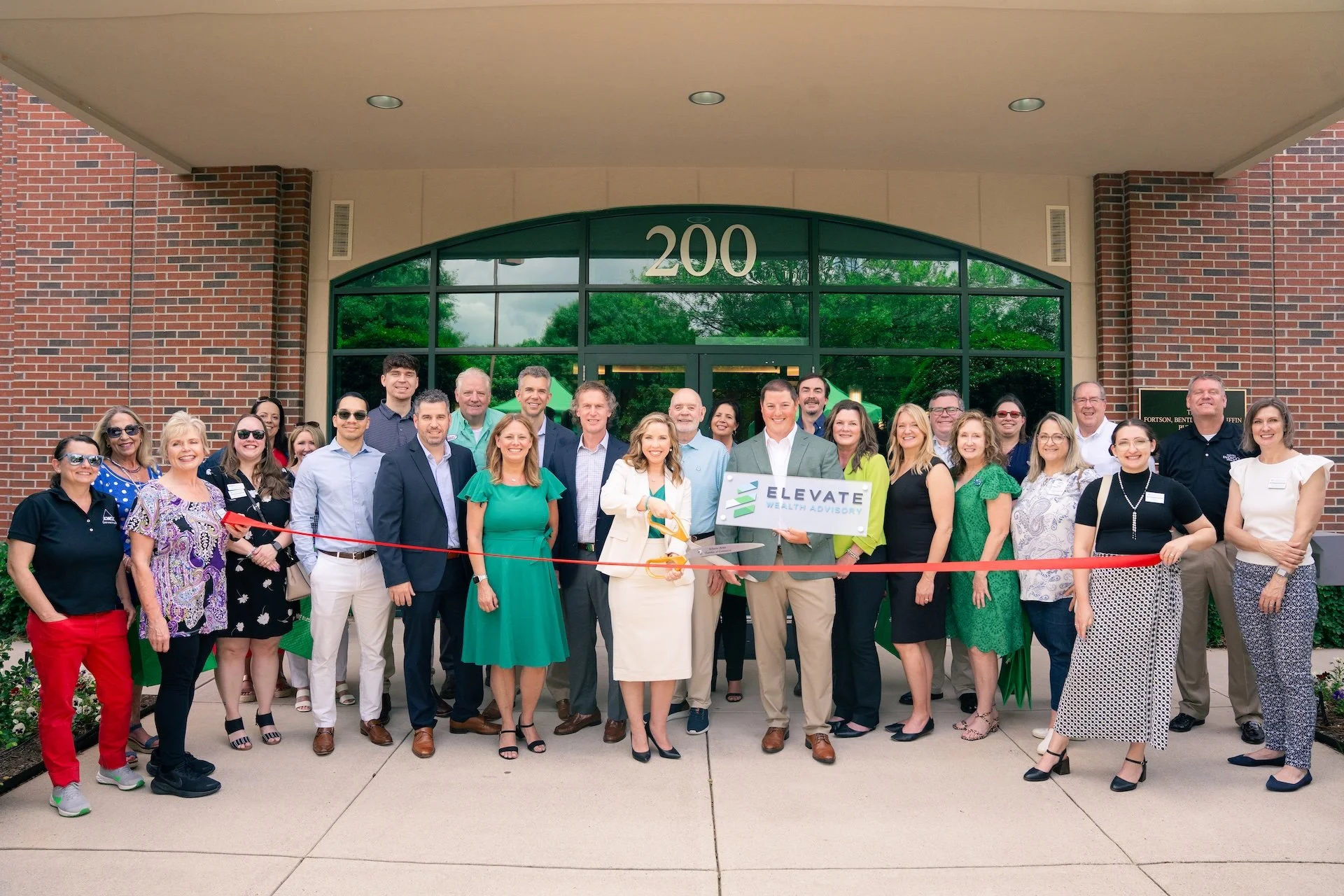 Group of people celebrating the opening of a new office building with a ribbon-cutting ceremony in front of a brick building with large glass windows and the number 200 above the entrance.