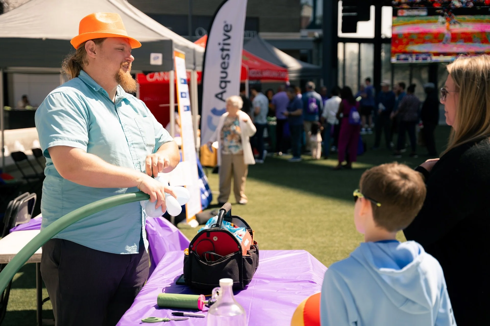 A man in a light blue shirt and orange hat speaking to a woman and a child at an outdoor event with tents and people in the background.