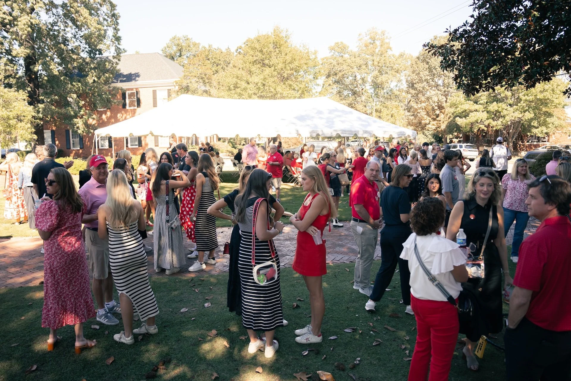 A diverse group of people at an outdoor gathering with a large white tent, in a park-like setting on a sunny day, with trees, grass, and brick pathways.