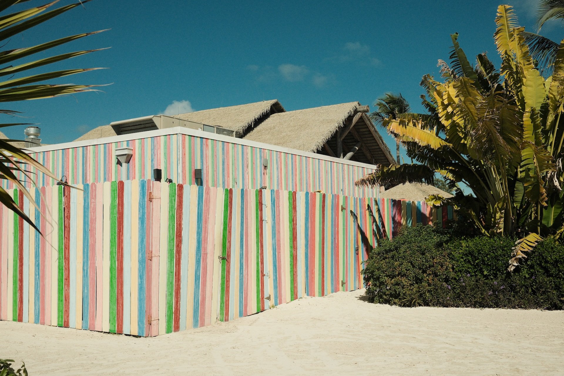 A beachside building with a colorful striped fence and a bamboo roof, surrounded by tropical plants and palm trees, under a blue sky.
