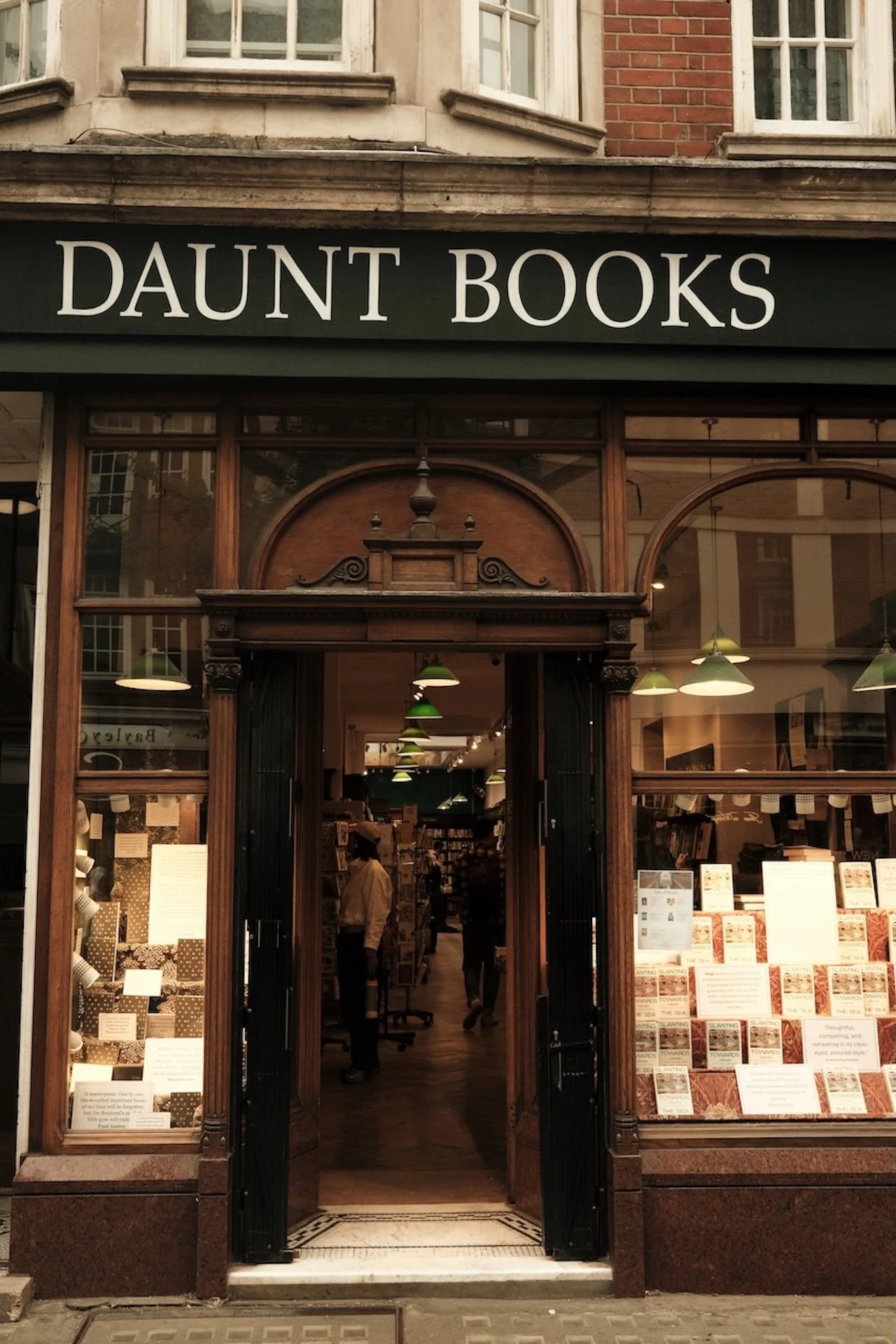 Storefront with sign 'Daunt Books' in black and white. Wooden door open, inside visible with bookshelves, green pendant lights, and customers.