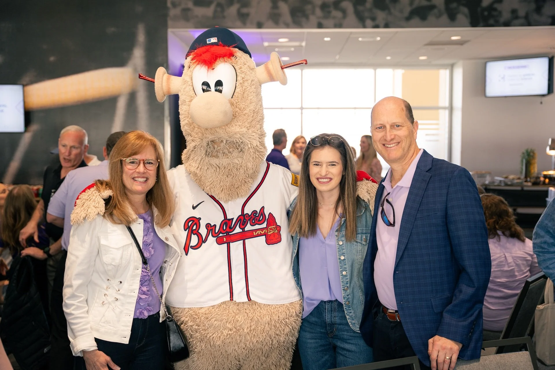 Four people standing together indoors, including a person in a Braves mascot costume. The mascot has a large furry body, a baseball cap, and a jersey with 'Braves' written on it. The group appears happy, smiling at the camera.