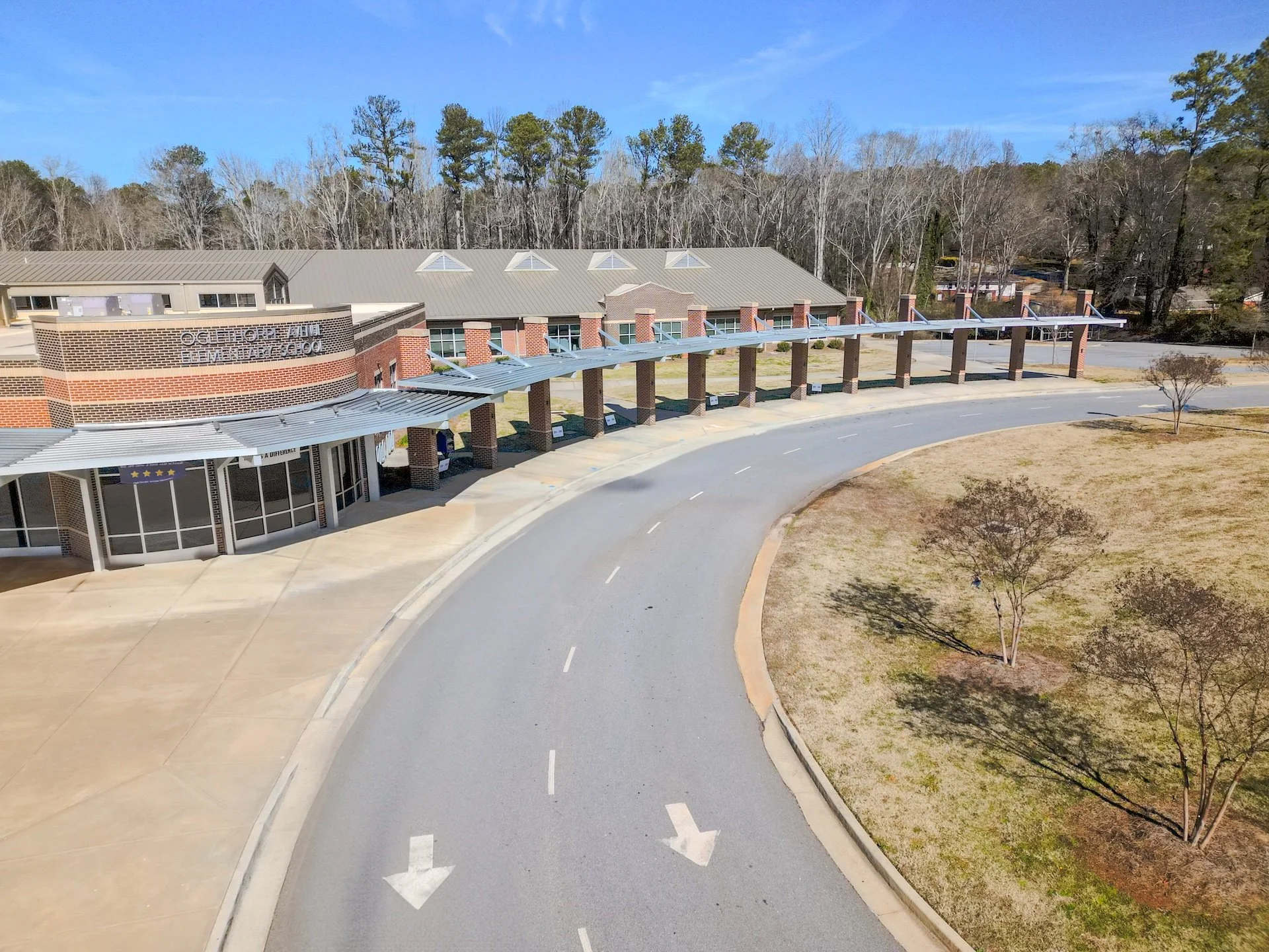 Aerial view of a school entrance with a curved road, brick and glass building, metal awning, and trees.