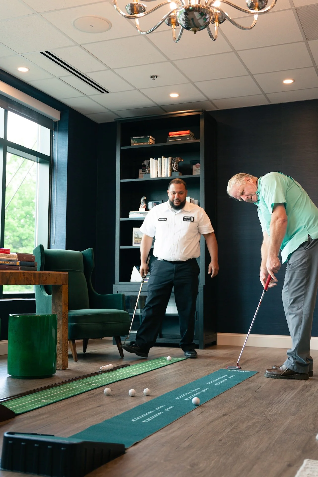 Two men playing miniature golf indoors, one holding a putter and the other observing. There is a green putting mat and several golf balls on the floor.