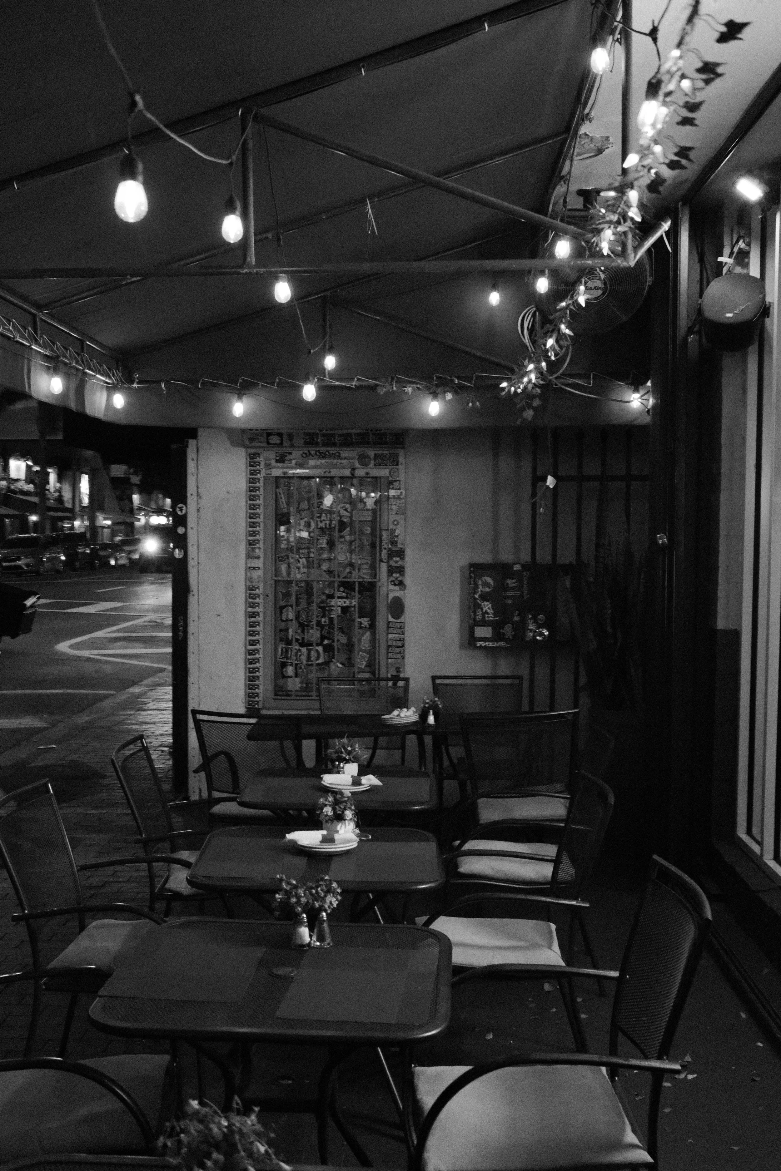 Outdoor patio of a restaurant with tables, chairs, small flower arrangements, string lights, and a vending machine inside.