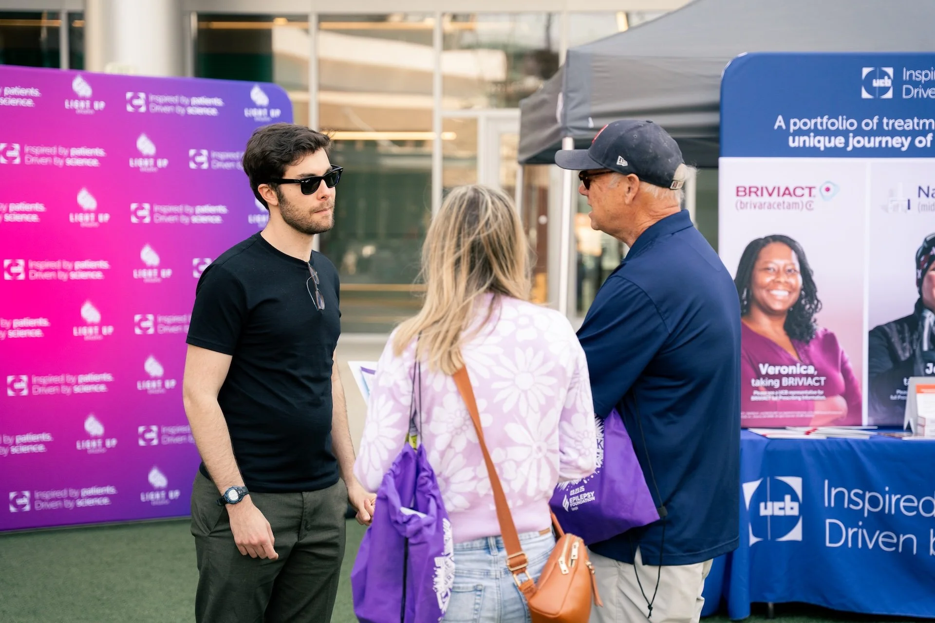 A man wearing sunglasses, a woman with a purple bag, and an older man with a cap are talking at an outdoor health event booth with banners and tables.