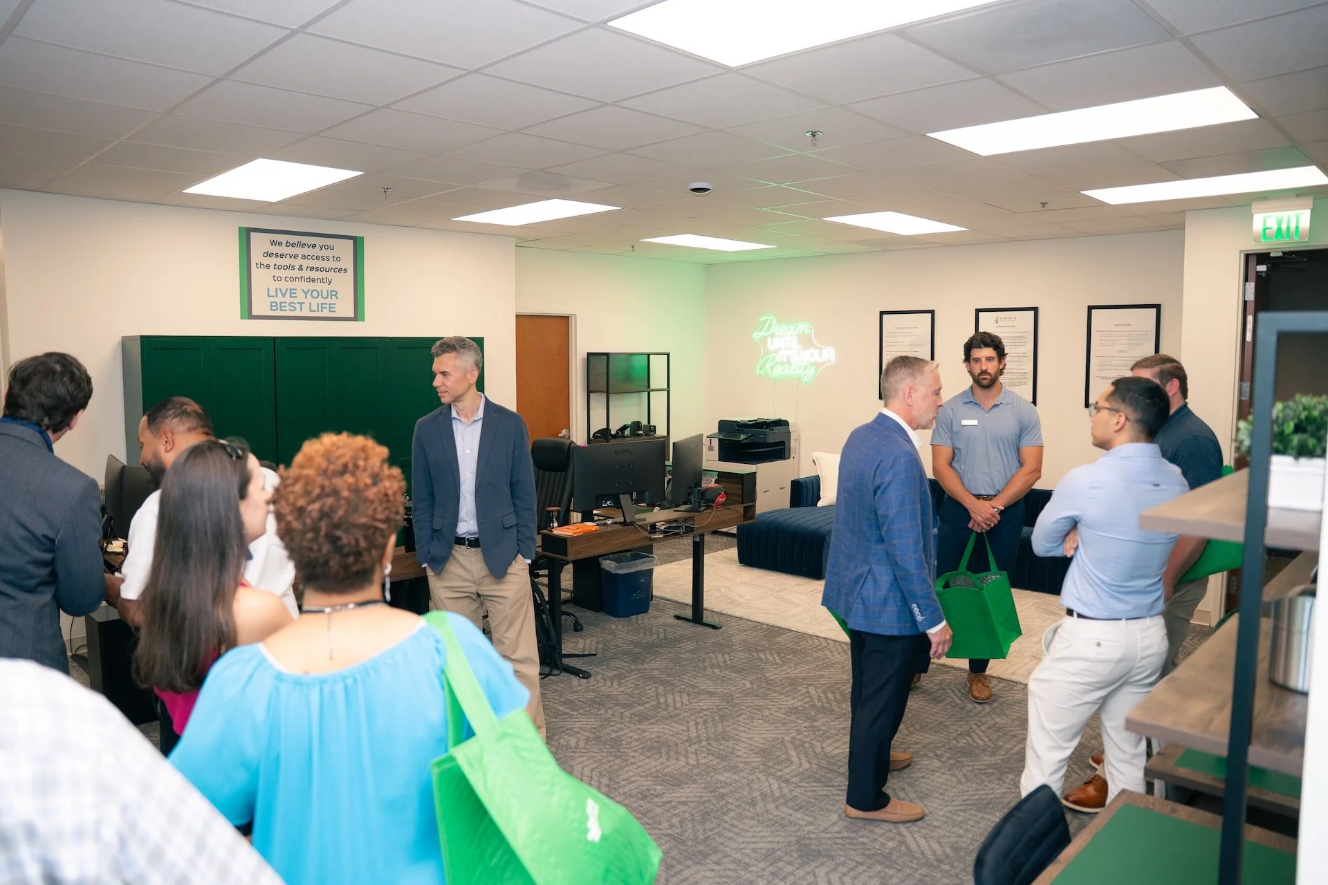 A group of professionals socializing in an office lounge area with white walls, hanging framed documents, a neon sign that says 'Dream, Live, Grow,' and a sign on the wall that reads 'We believe you deserve access to the tools & resources to confiden