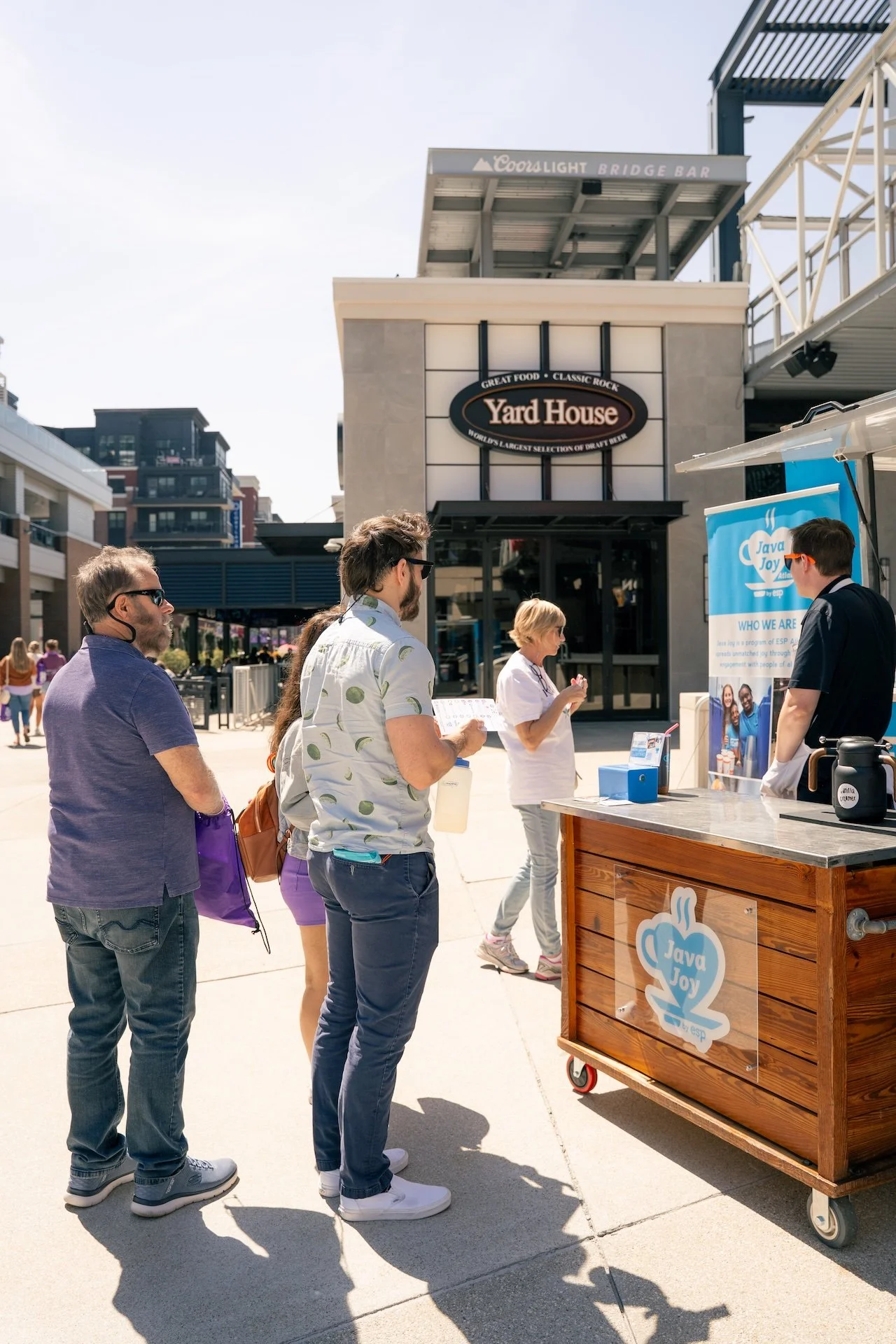 People standing in line at a Java Joy coffee stand outdoors, with a Yard House restaurant in the background.