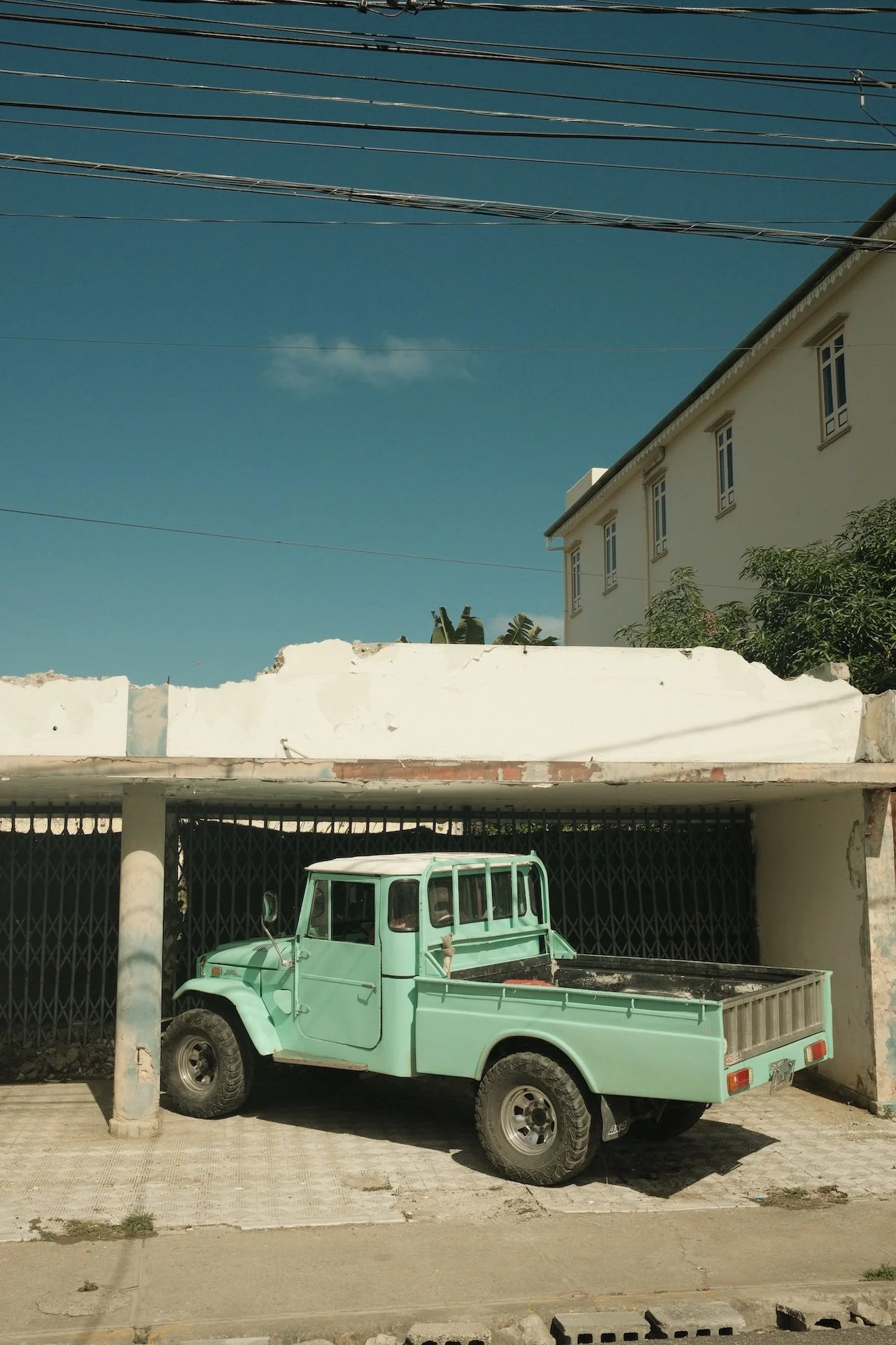 A mint green vintage pickup truck parked under a white concrete structure on a sidewalk, with a dark metal gate behind it and a white building with windows and trees in the background under a clear blue sky.