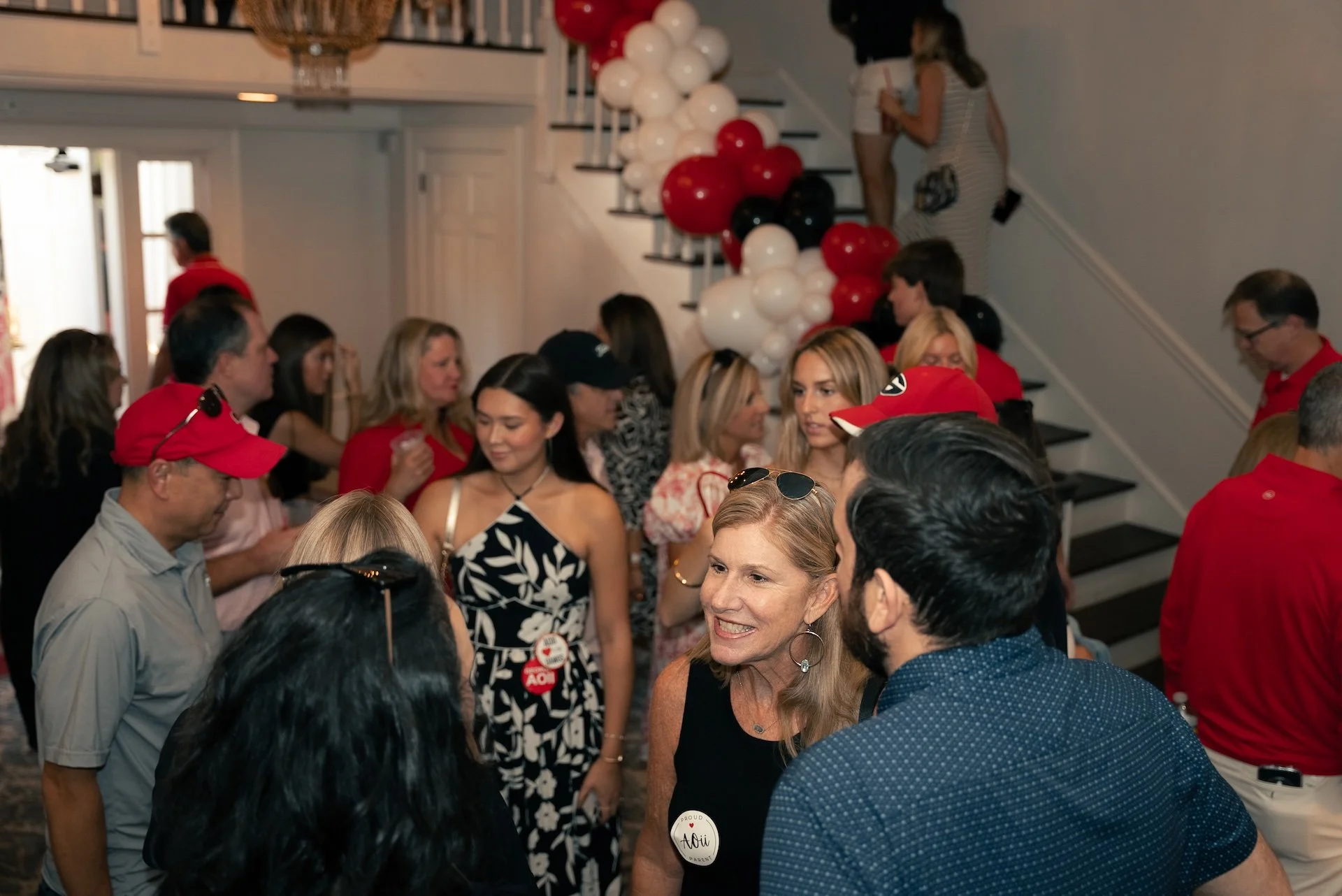 People gathered at a social event in a house, with red, white, and black balloons decorating stairs, and attendees wearing casual and semi-formal clothing, including some in red caps and shirts.