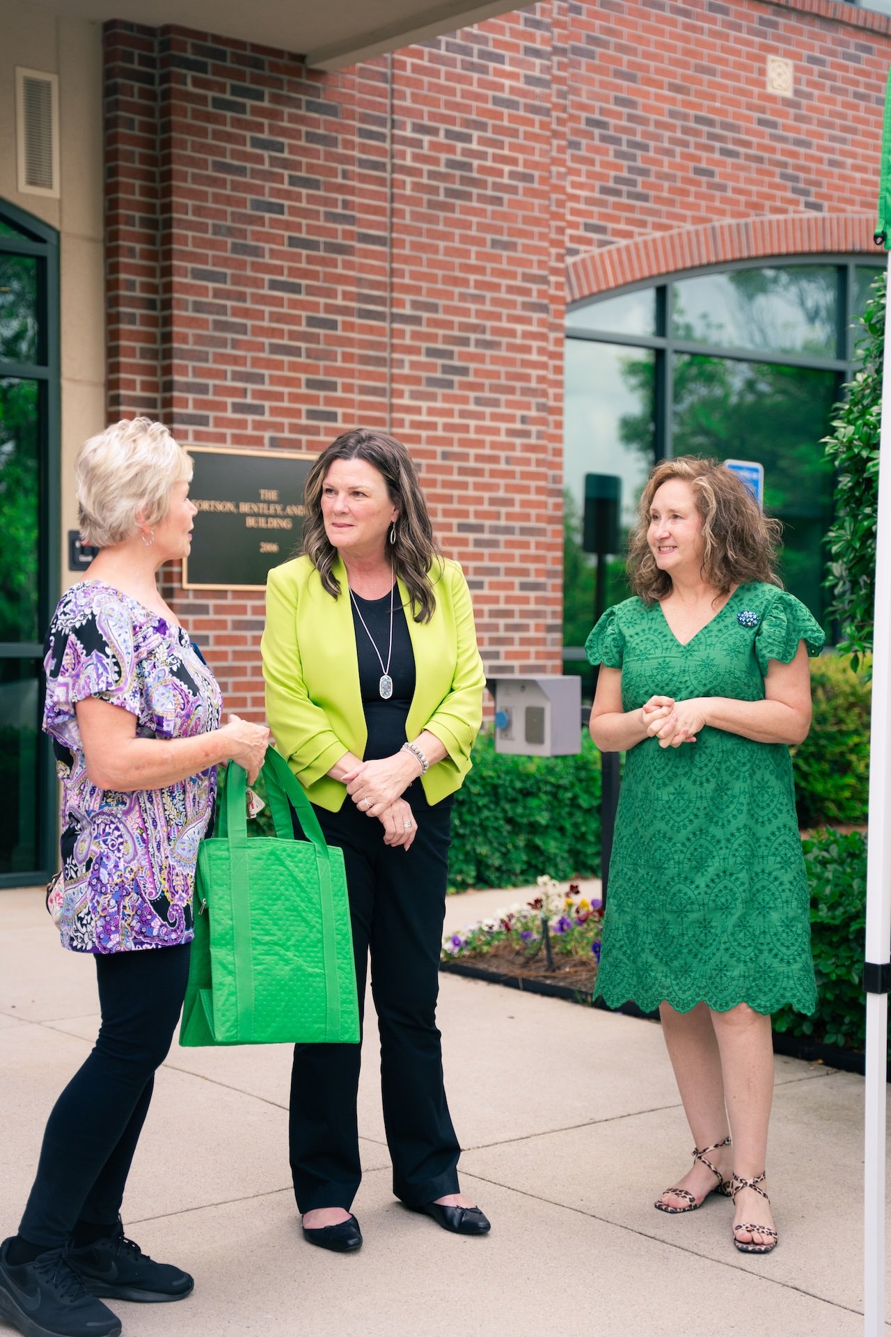 Three women standing outside near a brick building engaged in conversation. One woman has short blonde hair, patterned purple blouse, black pants, and holds a green tote bag. The second woman has long dark hair, green blazer, black pants, and is wear