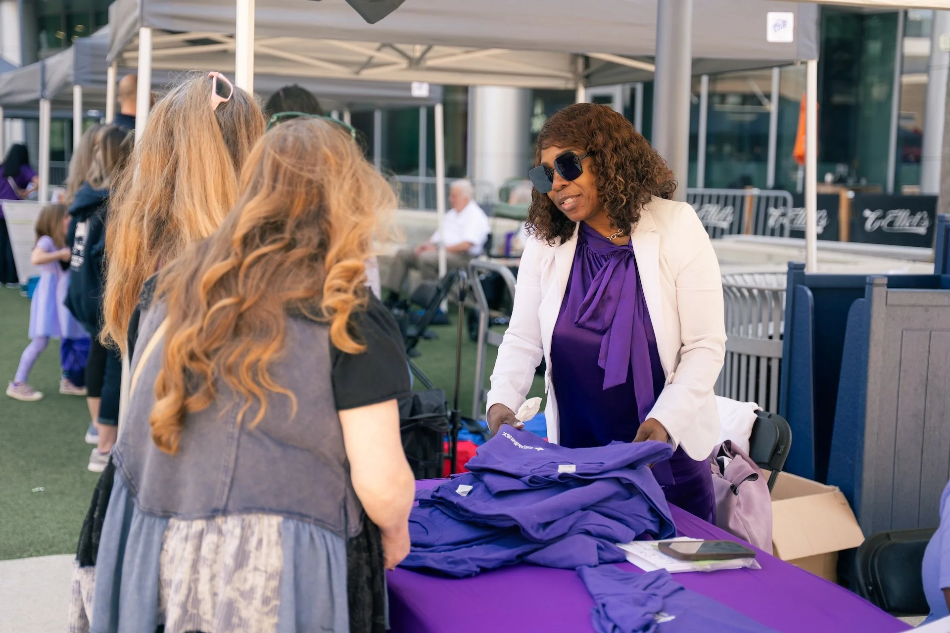 A woman in sunglasses and a white blazer showing purple t-shirts to two women at an outdoor booth with a purple tablecloth.