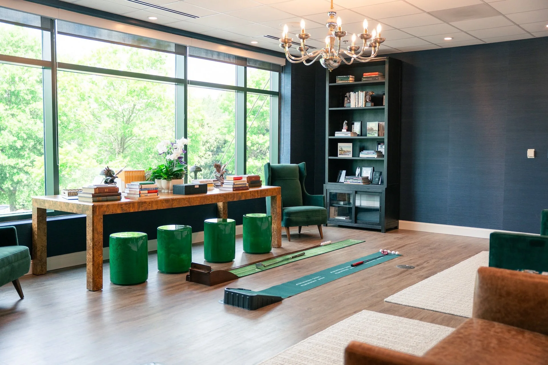 An office or lounge with large windows, a wooden desk, green stools, a green high-back chair, a bookshelf, and a chandelier.