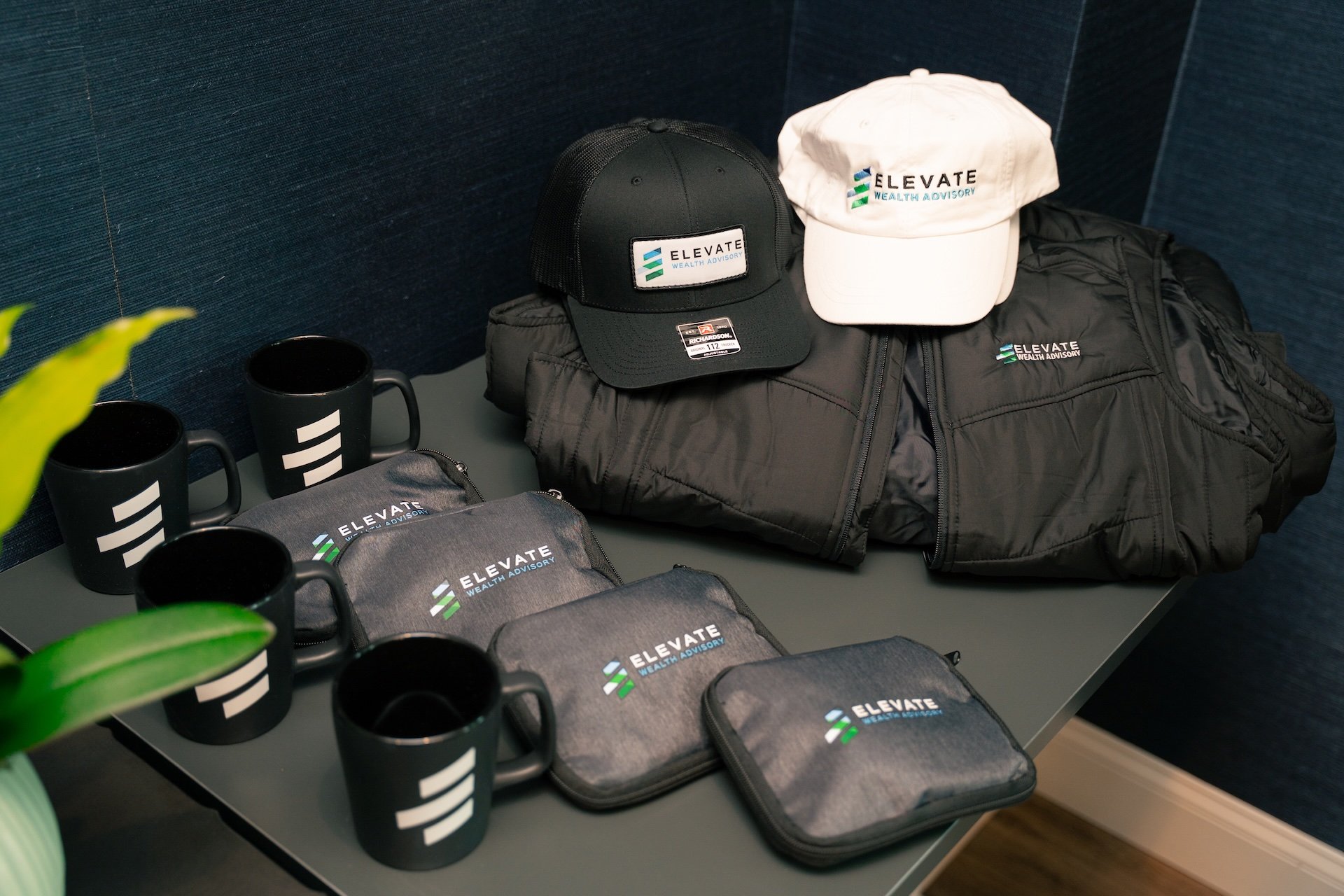 Branded merchandise including black and white caps, black jackets, mugs, and pouches with the Elevate Wealth Advisory logo on a table.