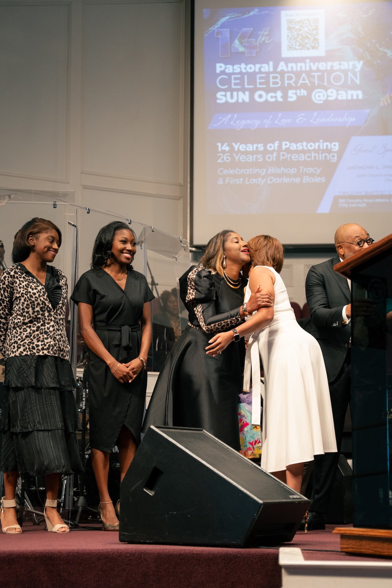 A group of people on stage during a celebration event, with a large screen in the background displaying details about a pastoral anniversary and celebration.