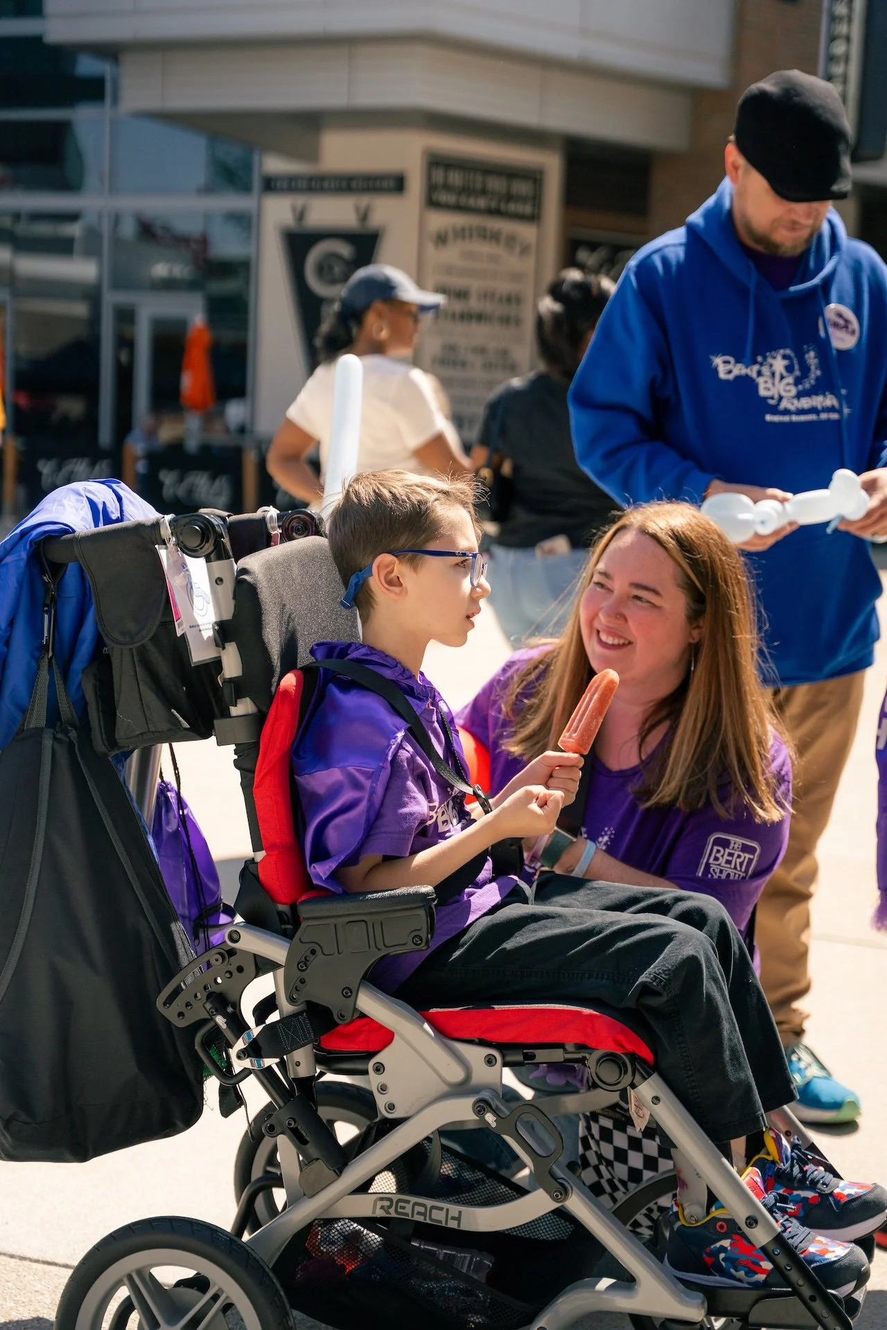 A young boy in a wheelchair holding a popsicle and talking to a woman in a purple shirt. There are people in the background at an outdoor event.