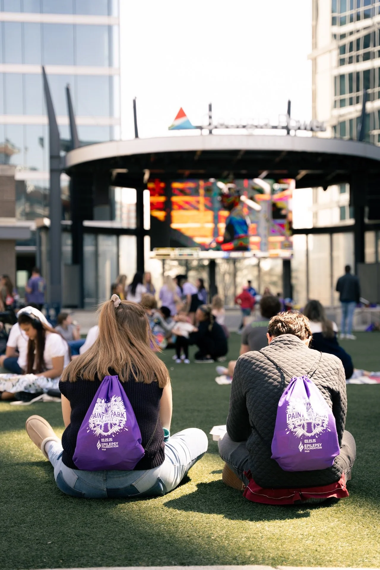 Two people with purple backpacks sitting on a grassy area at an outdoor event, facing a stage with a large screen, in an urban setting with modern glass buildings.