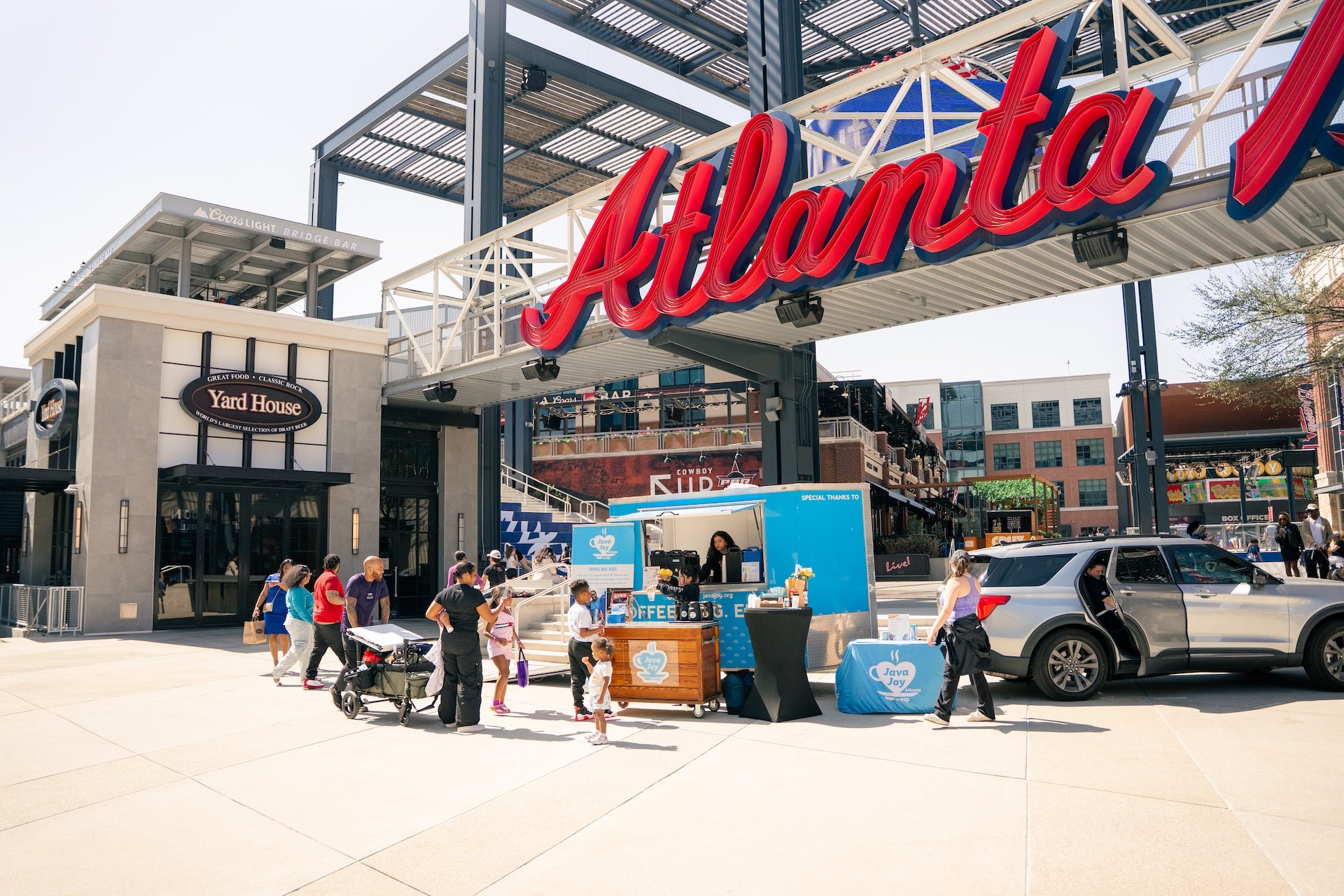 People gather around a coffee stand labeled 'Java Joy' outside the Atlanta amusement park, with the park's red 'Atlanta' sign overhead and various buildings and attractions in the background, on a sunny day.