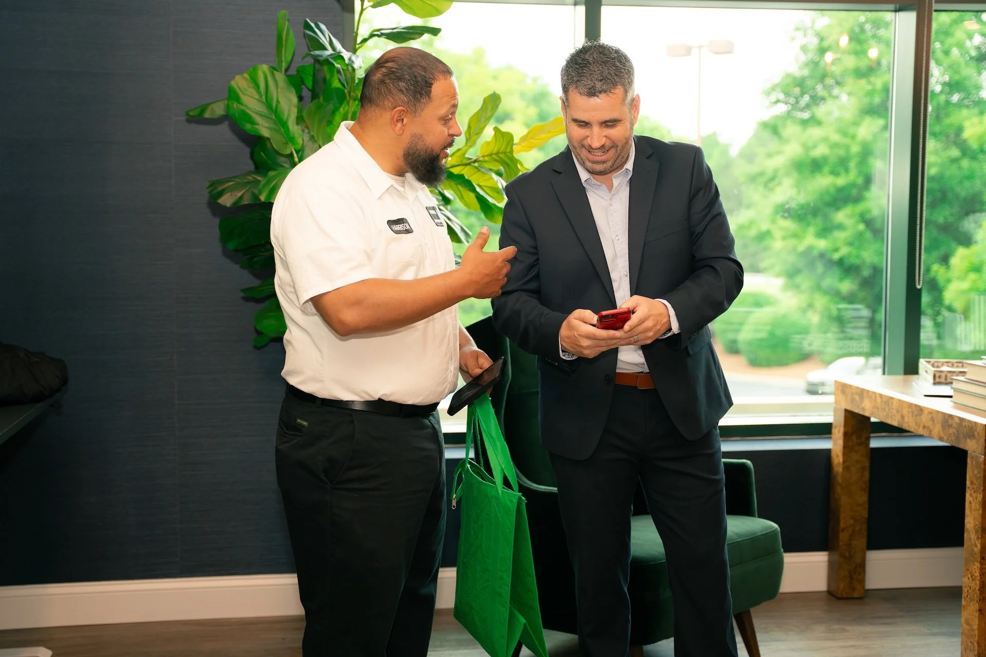 Two men, one in a white shirt with a name tag and the other in a dark suit, are standing in a modern office. They are smiling and looking at a red smartphone that the man in the suit is holding. The man in the white shirt is also holding a black fold