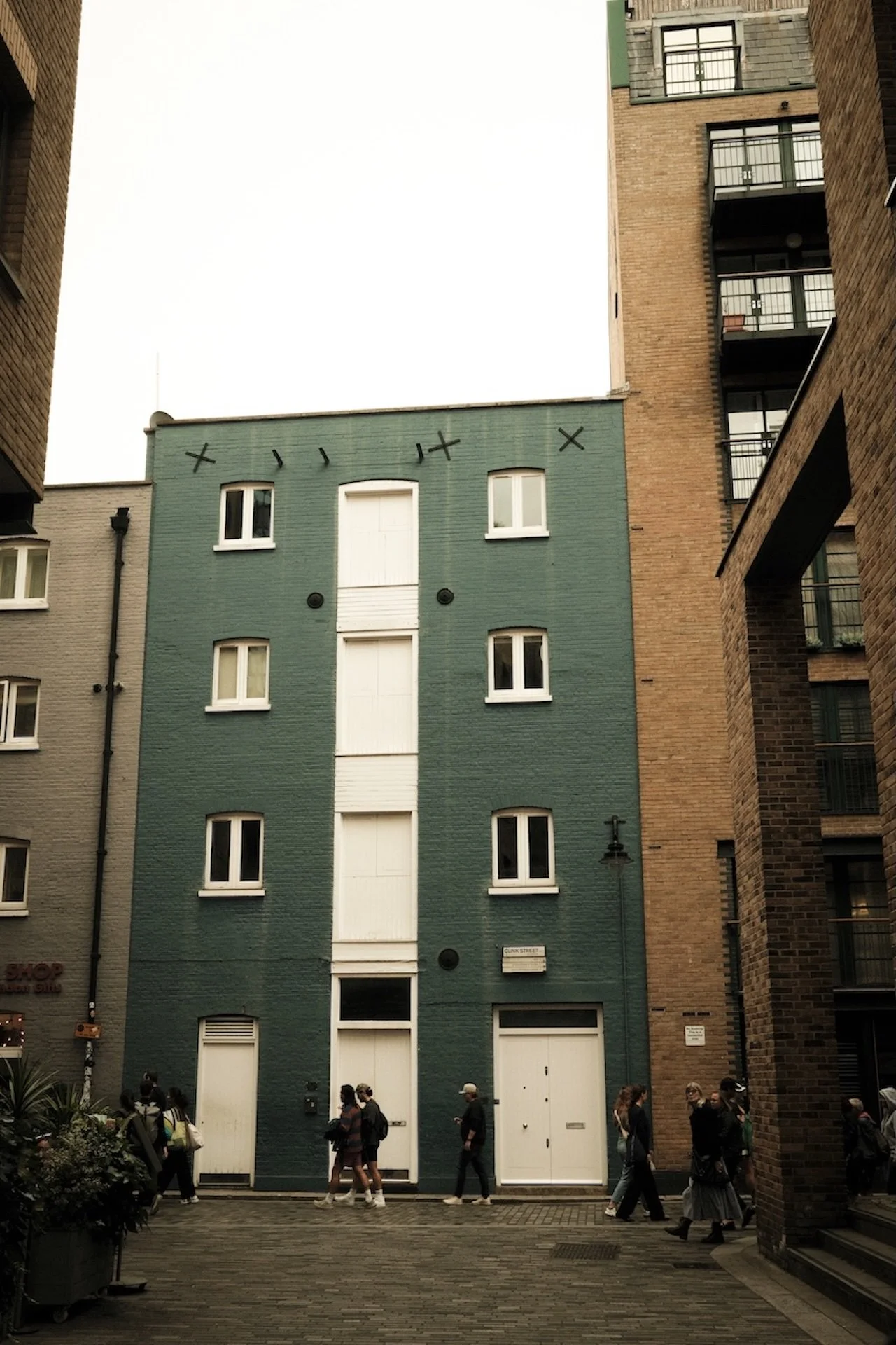 A tall green building with white windows, situated between two taller brick buildings, with people walking on a cobblestone street in front.