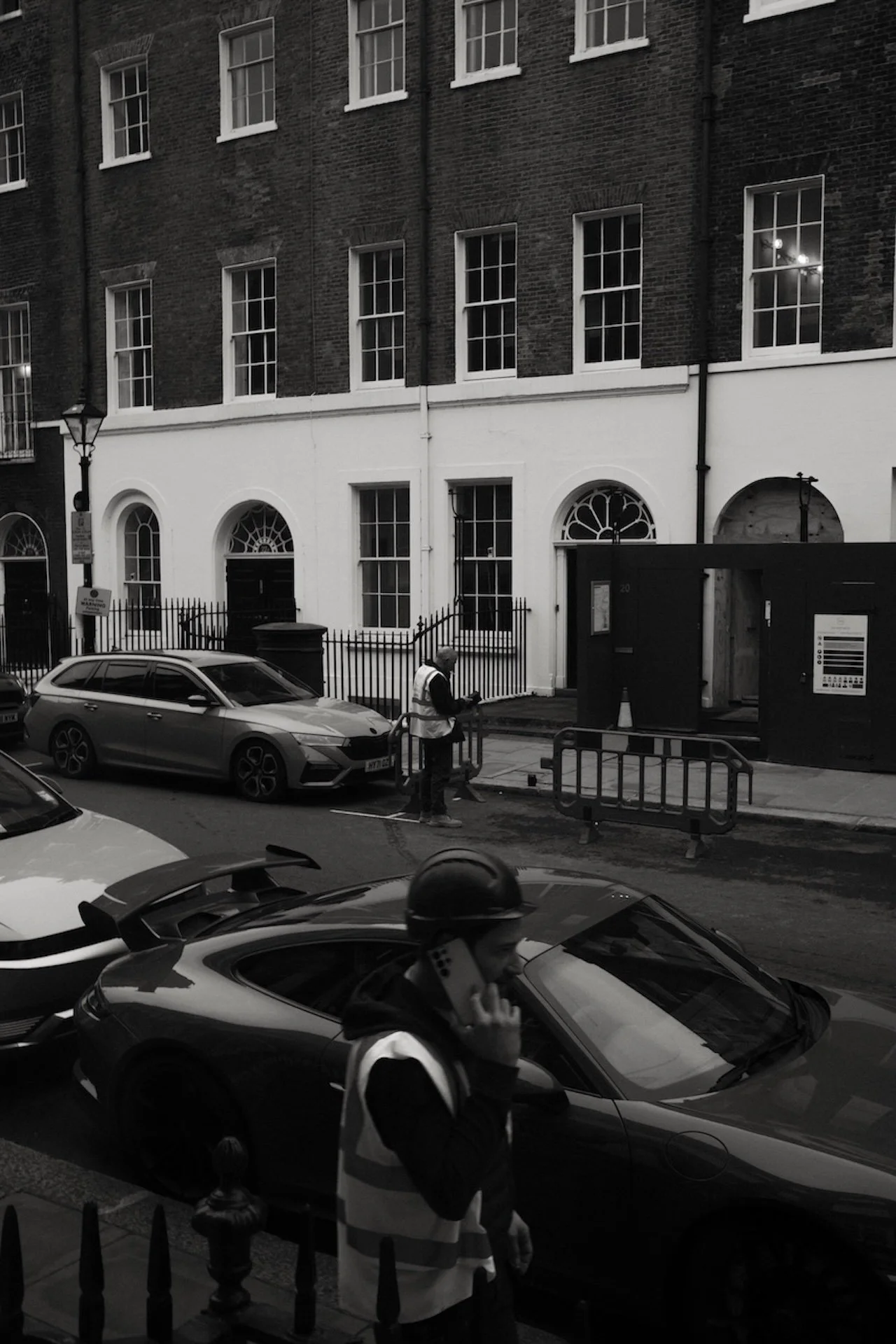 A street scene in black and white with parked cars, a person wearing a helmet and talking on a phone, another person working on the sidewalk, and a row of tall buildings with large windows.