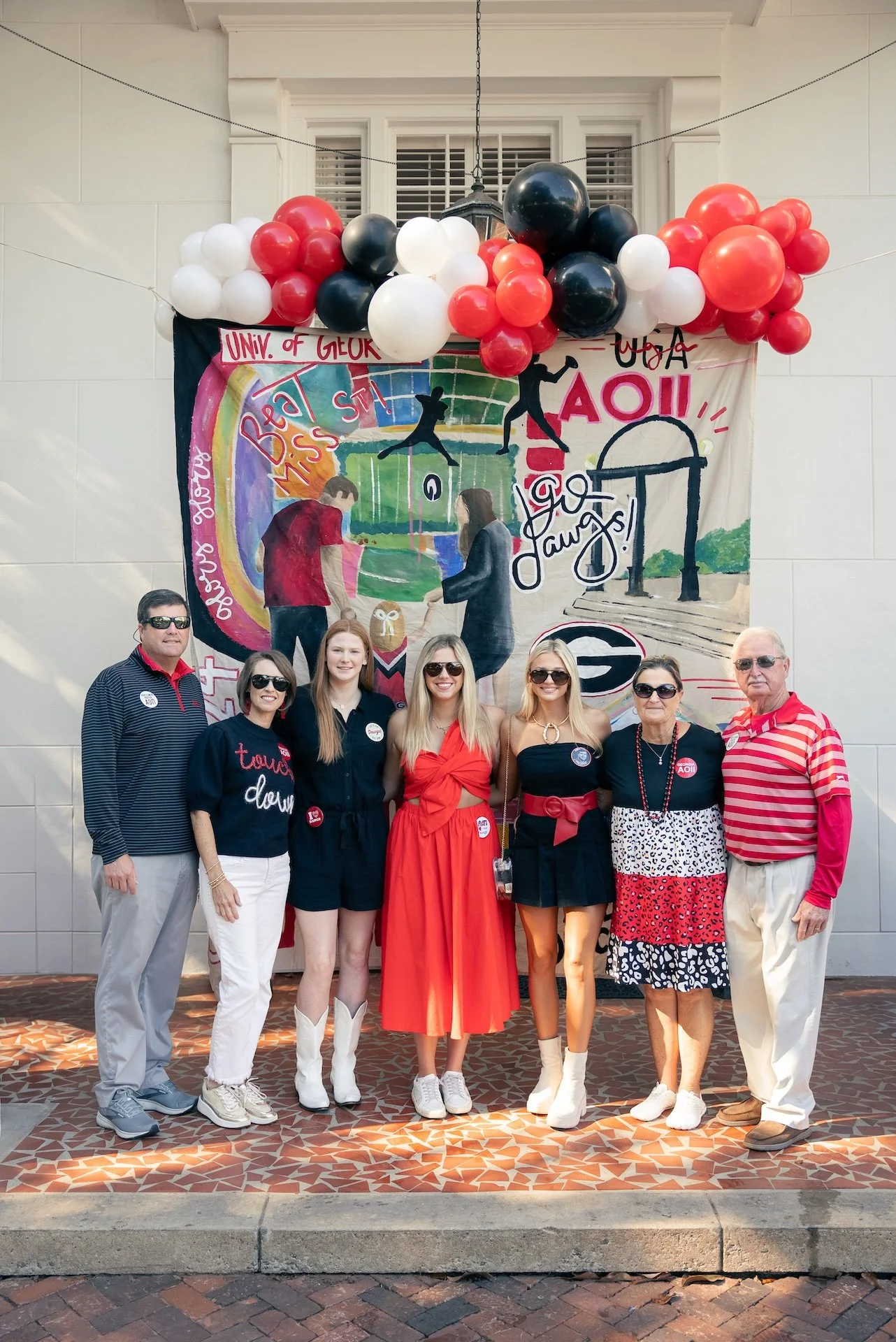 Group of seven people standing in front of a colorful mural and red, black, and white balloons, celebrating an event related to the University of Georgia.